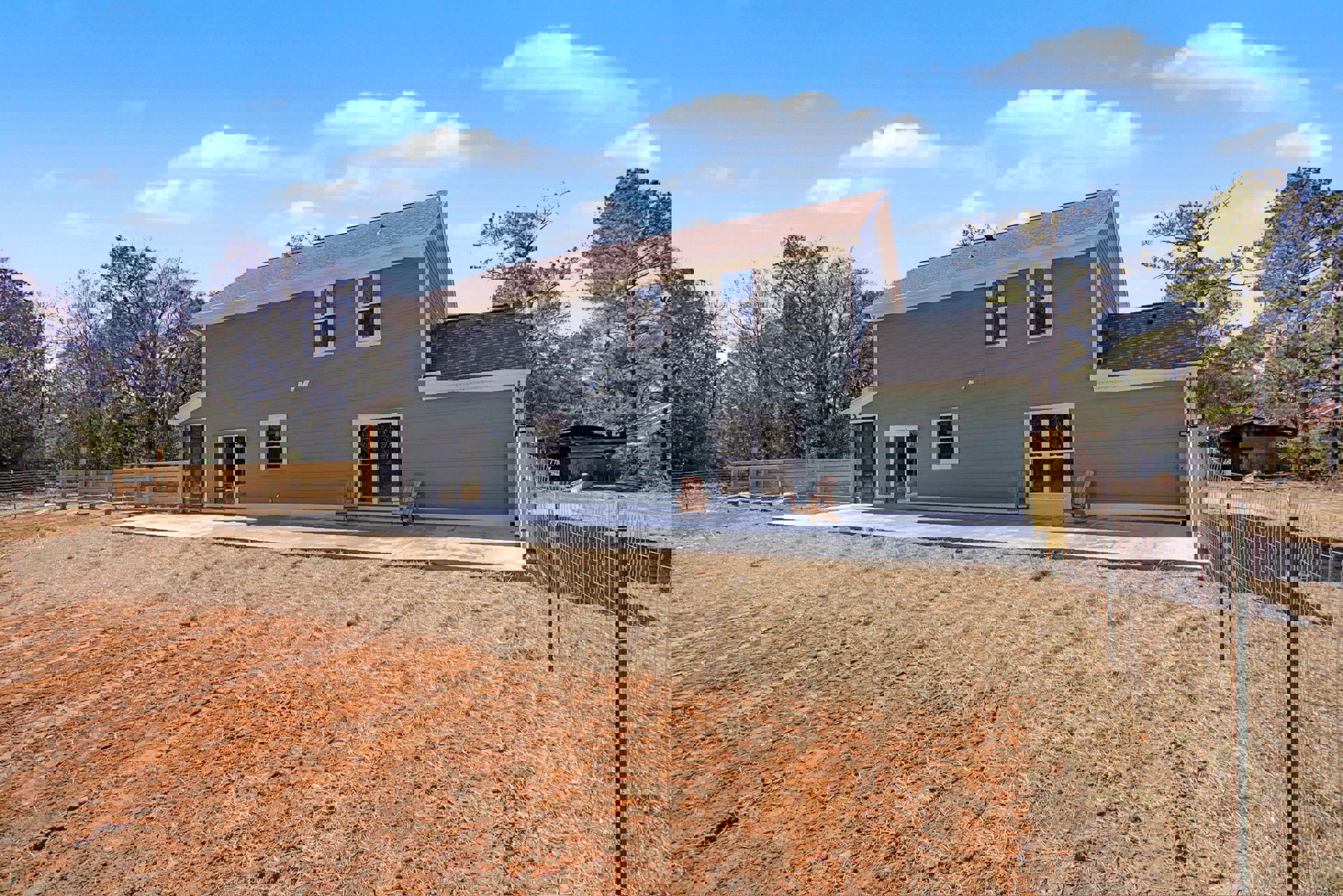 Two-story home with white-framed windows, blue glass accents, wooden porch chair, fenced yard, green lawn, and mature trees under a partly cloudy sky.