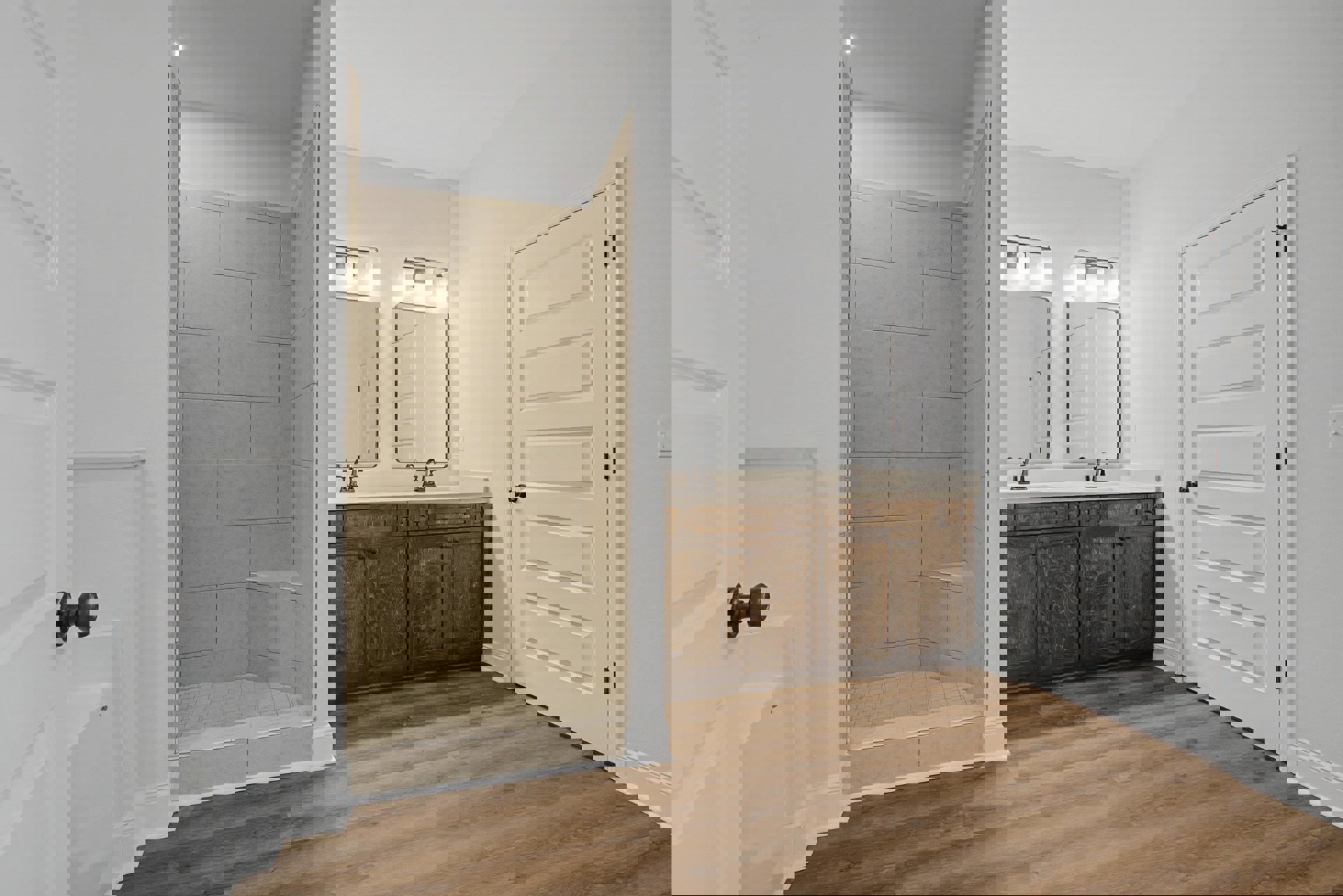 Bathroom featuring a glass-enclosed shower with tile wall, double sinks set in white cabinets with black trim, wood flooring, white baseboards, and a white door with black handle