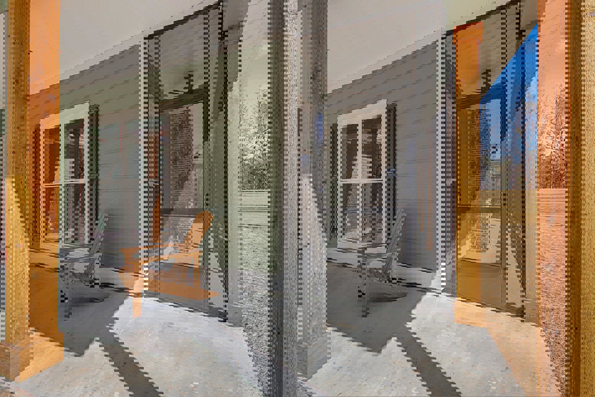 Wooden porch with a single chair beside a screen door, glass-paned window, light fixture on wood siding, and forest view through window