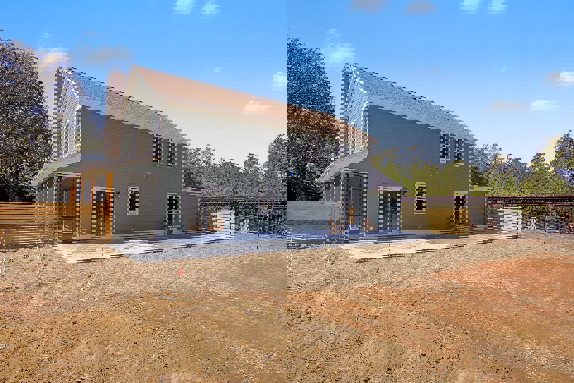 Modern home under construction featuring white-framed windows, open door, sliding glass door, lawn with a chair, and surrounding trees behind a wooden fence