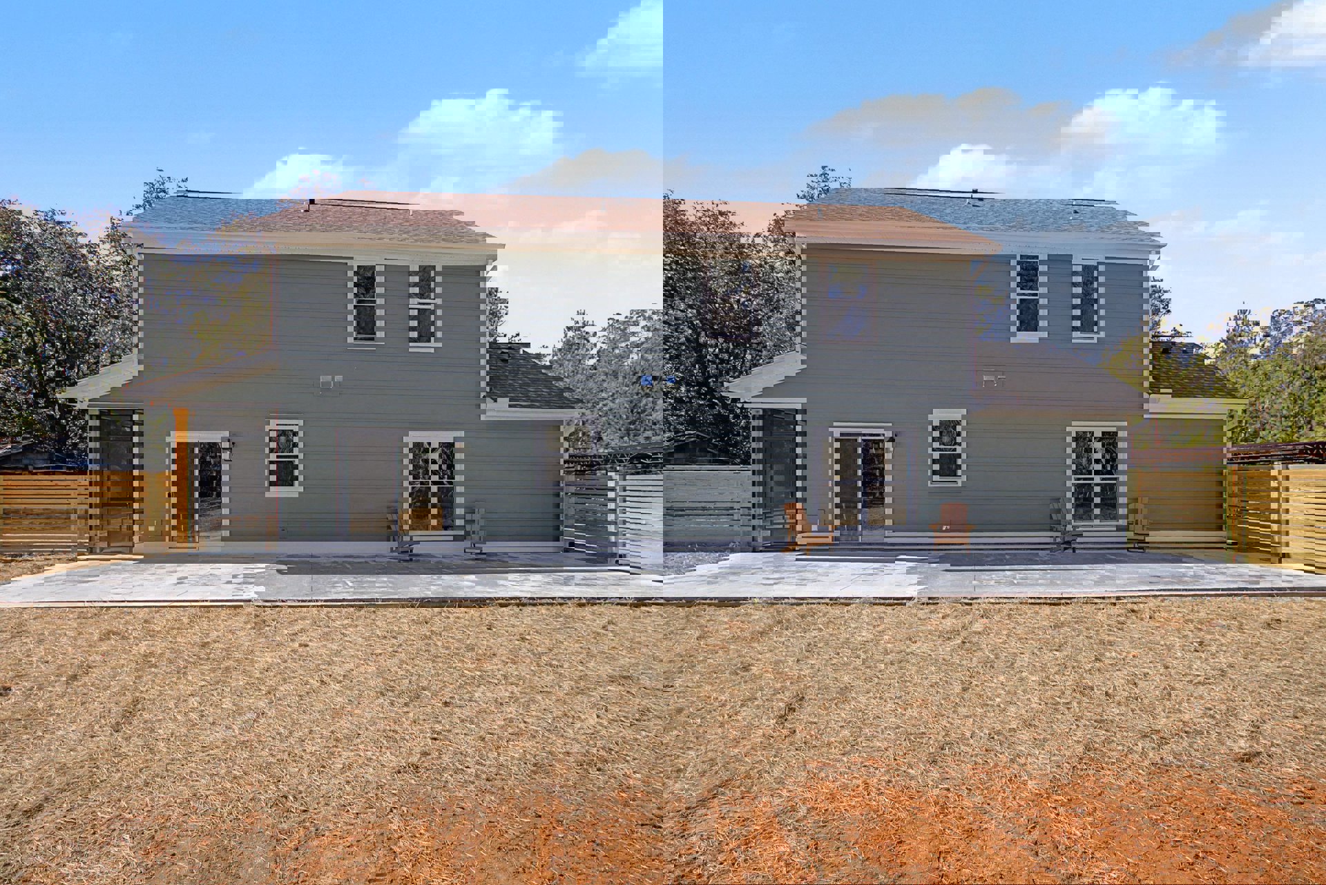Two-story house with white siding, large windows, green lawn, stone patio, and wooden outdoor chair near the entrance.