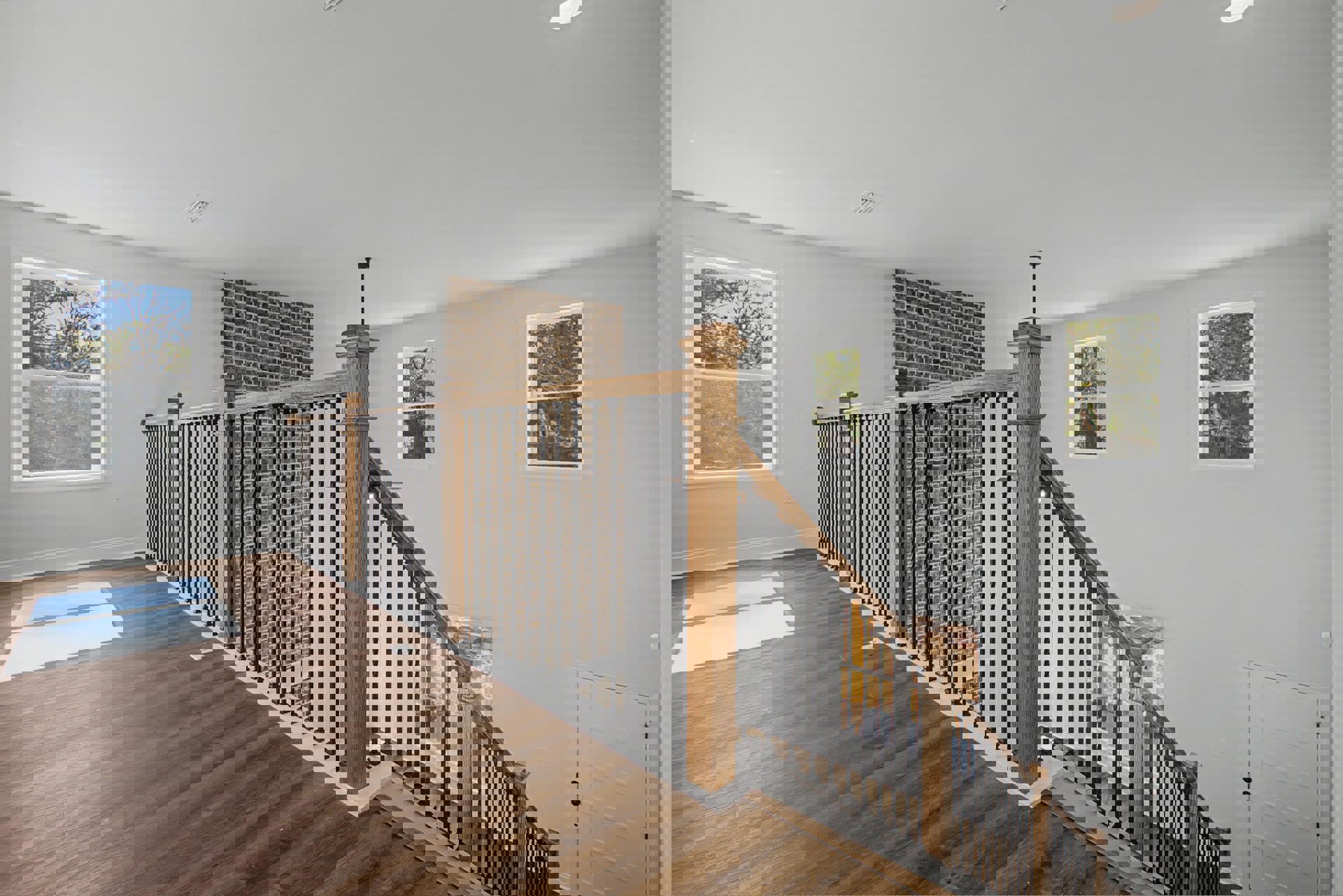 Wood staircase with metal railings beside an exposed brick wall, white door with black handles, large window overlooking trees, laminate flooring and plaster walls