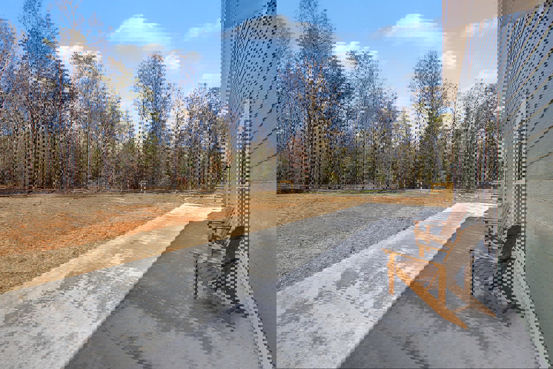 Wooden chairs arranged on a concrete patio deck, bordered by grass and dirt, with mature trees in the background under a partly cloudy sky.