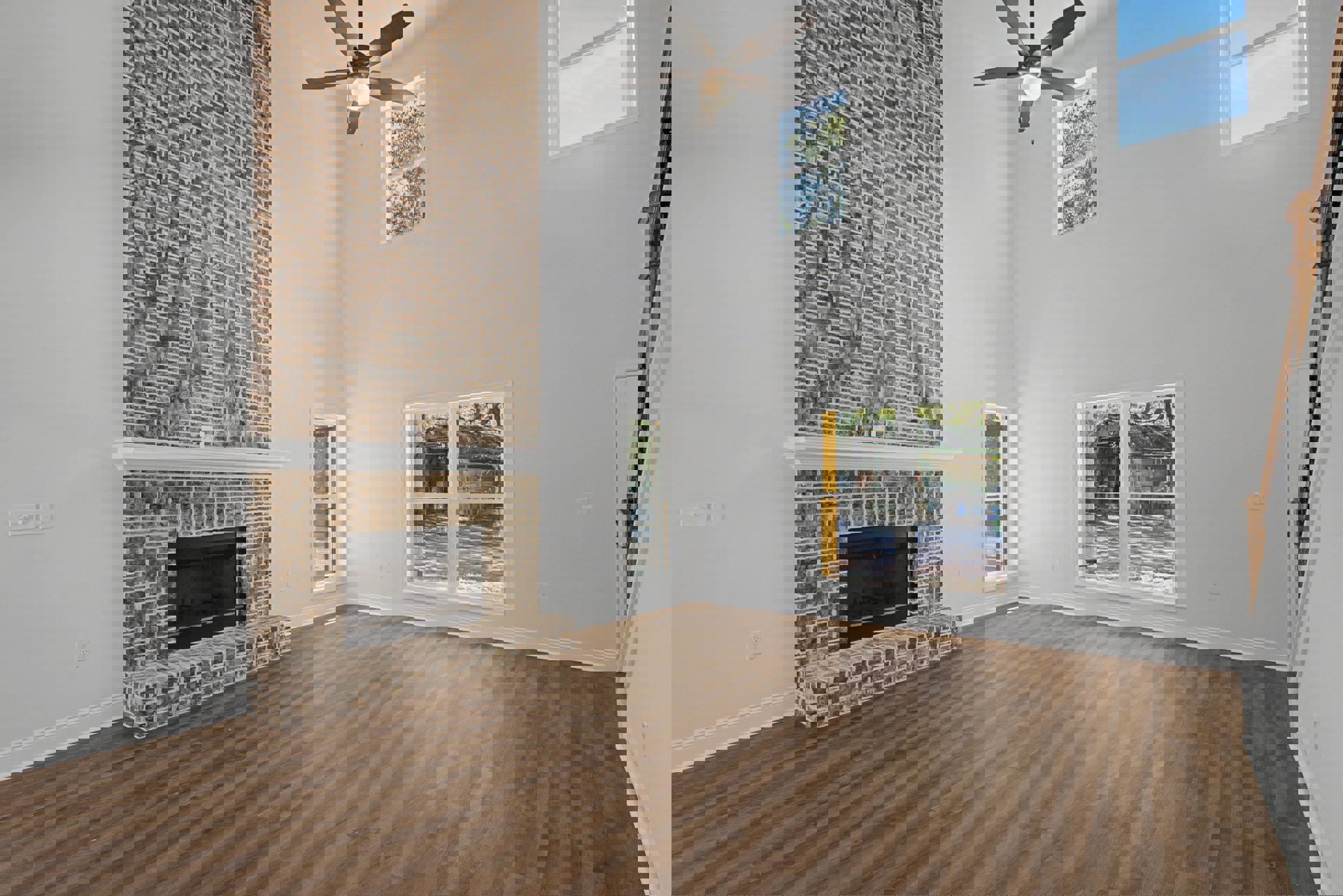 Living room with wood flooring, brick fireplace featuring a white mantle, ceiling fan with light, and window overlooking backyard shed