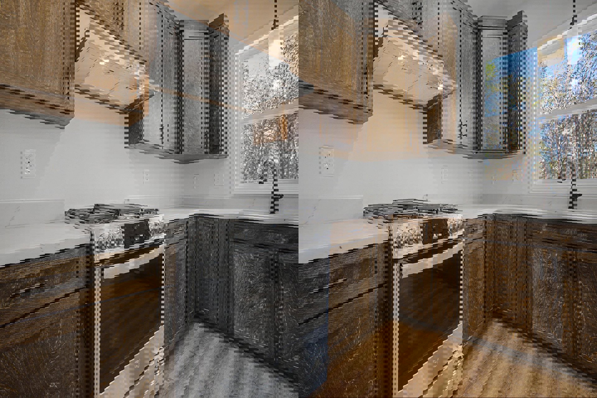 Kitchen with natural wood cabinets, stainless steel stove with glass door, matching range hood, stone countertop, window overlooking trees, pendant light fixture, and close-up