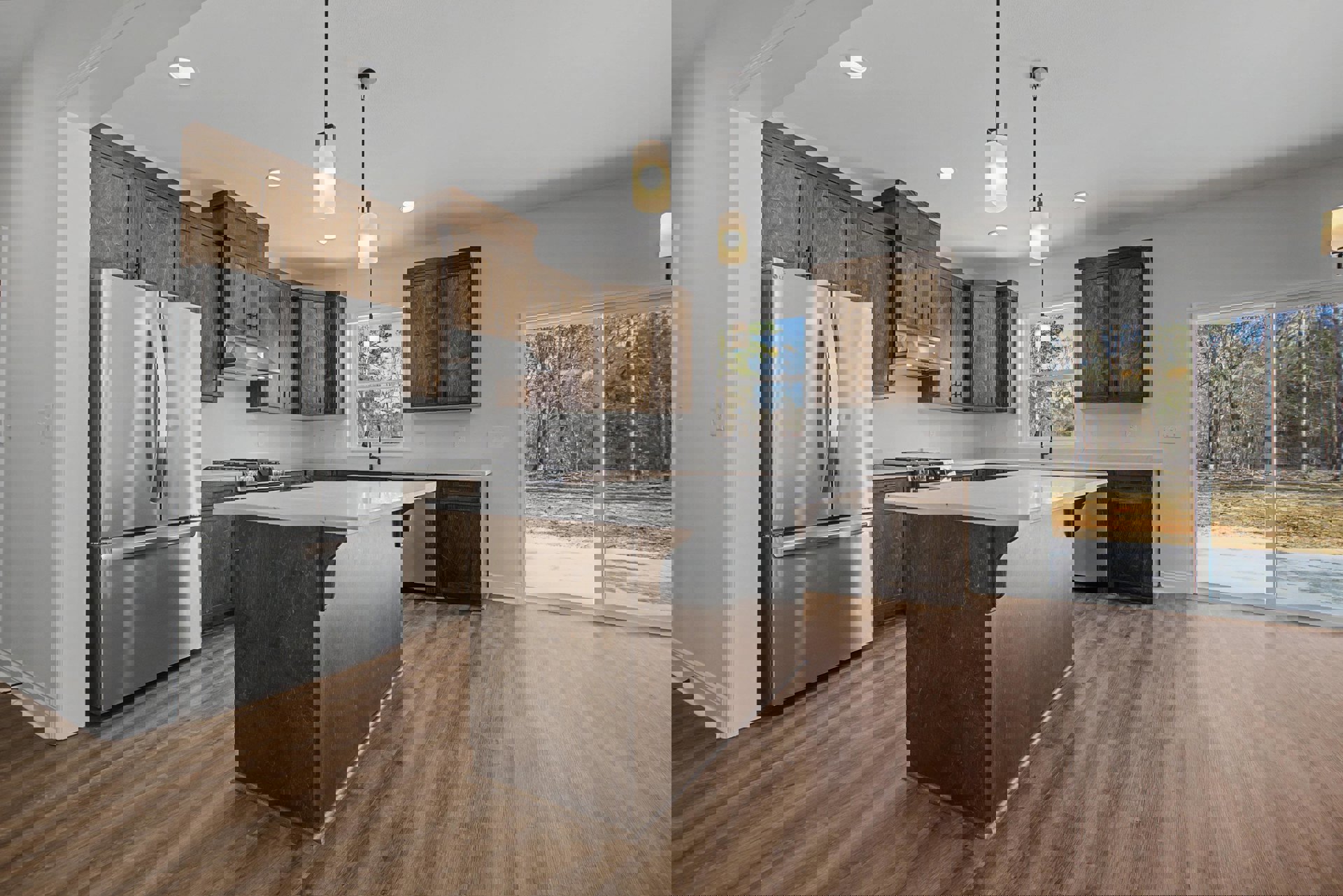 Kitchen with wide wood plank flooring, large central island with light stone countertop, stainless steel refrigerator, wood cabinets with metal handles, wall-mounted light fixture