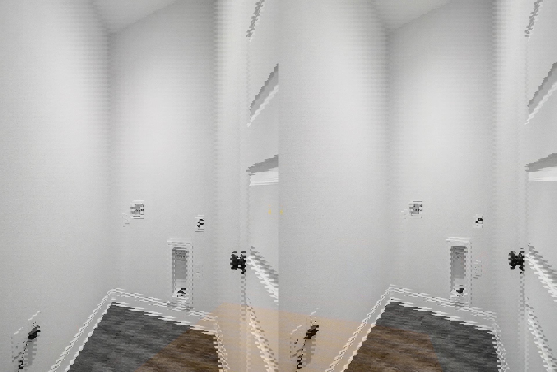 Bathroom with light wood flooring, white baseboards, white plaster walls, a white urinal with black lid, a white box with two valves, and a black rectangular object mounted on the