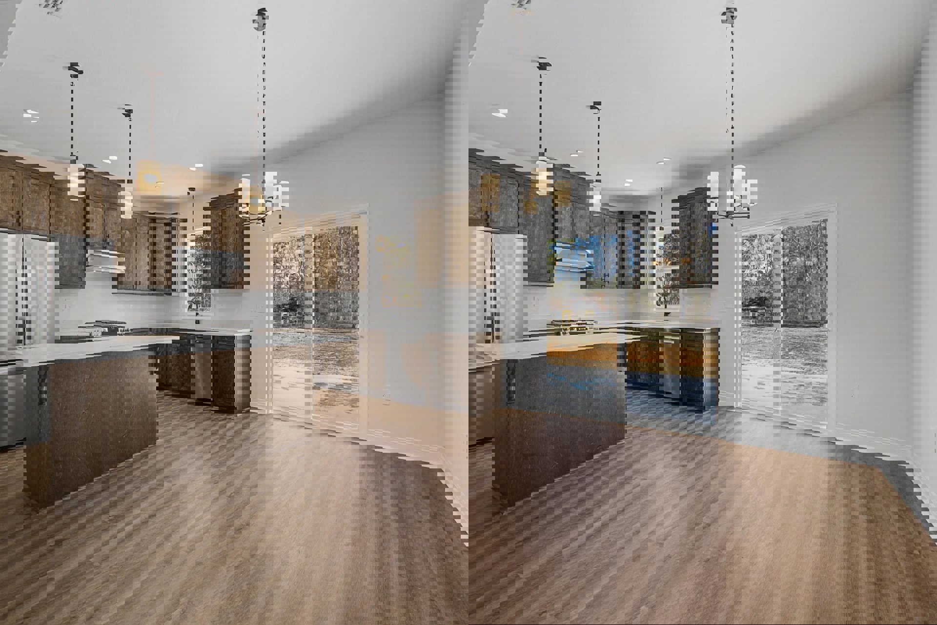 Kitchen with wide plank wood flooring, large window above sink, white cabinetry, stainless steel refrigerator, pendant light with white shade, sliding glass door opening to