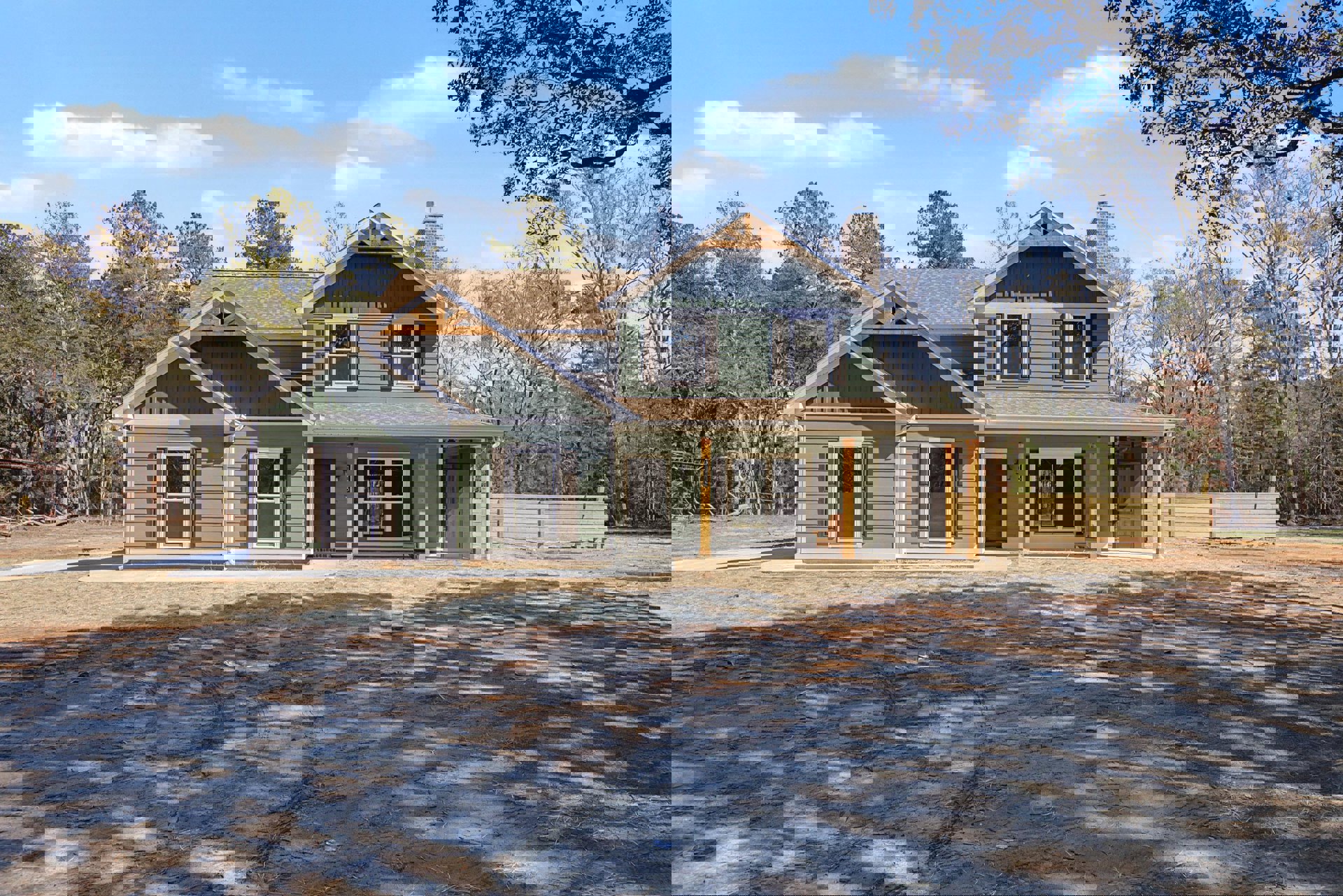 Modern home with white-framed windows, wood post porch, fenced yard, dirt driveway with tire tracks, and mature trees in the background