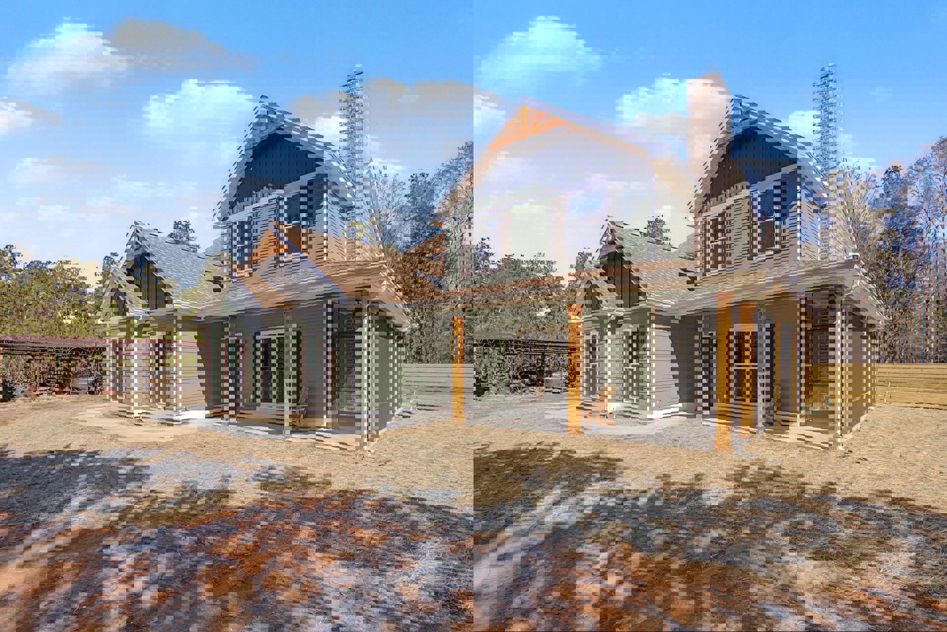 Two-story house with brick chimney, white-framed windows, covered porch with wooden chair, wooden bench in yard, tree casting shadow on lawn, metal roof structure in background.