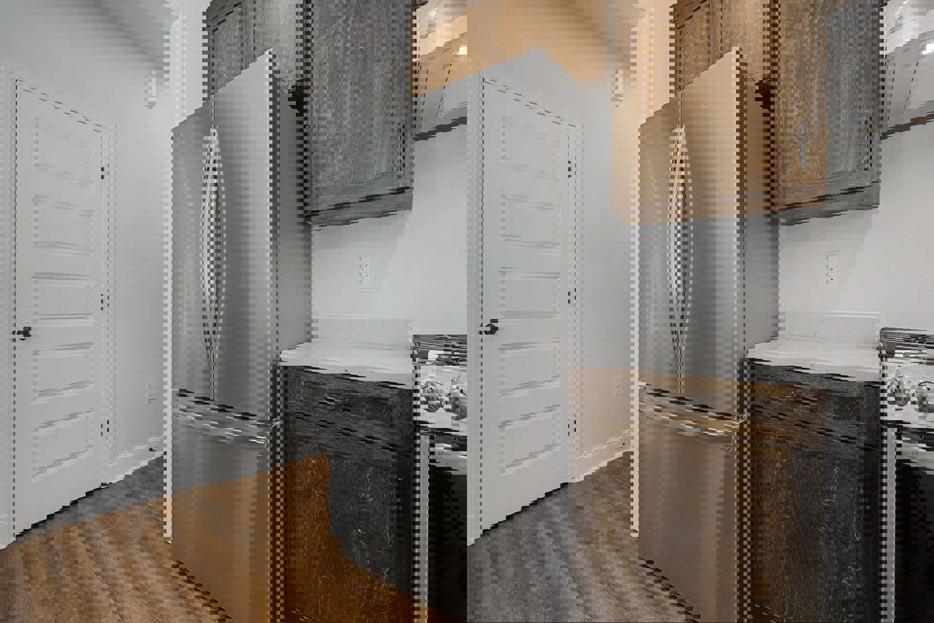 Stainless steel refrigerator beside white door with black handle, marble countertop atop wooden cabinets with black handles, close-up of stainless steel oven and electrical outlet