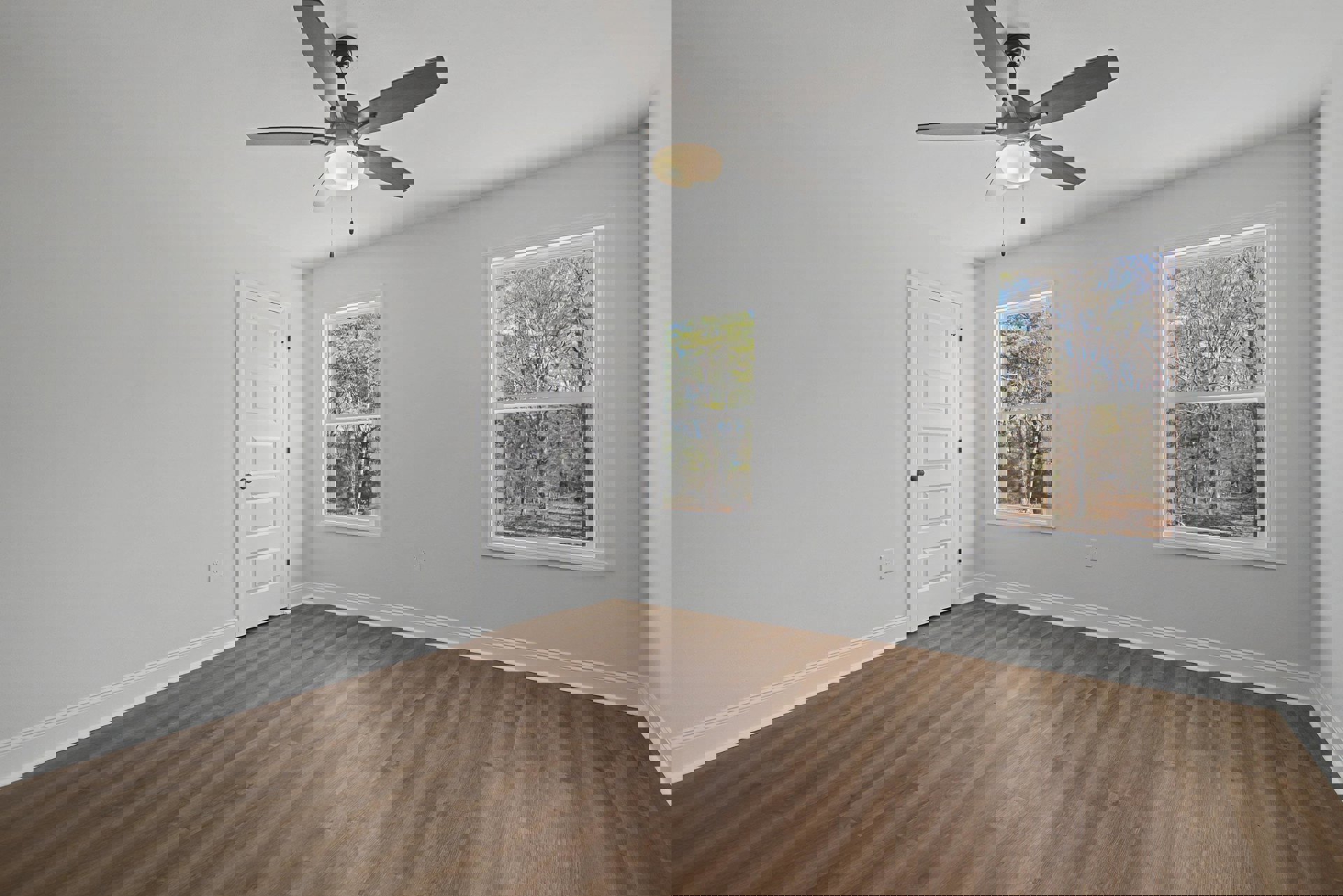 Ceiling fan with light fixture above wood flooring, white walls, large windows showing trees outside, white door with black hardware