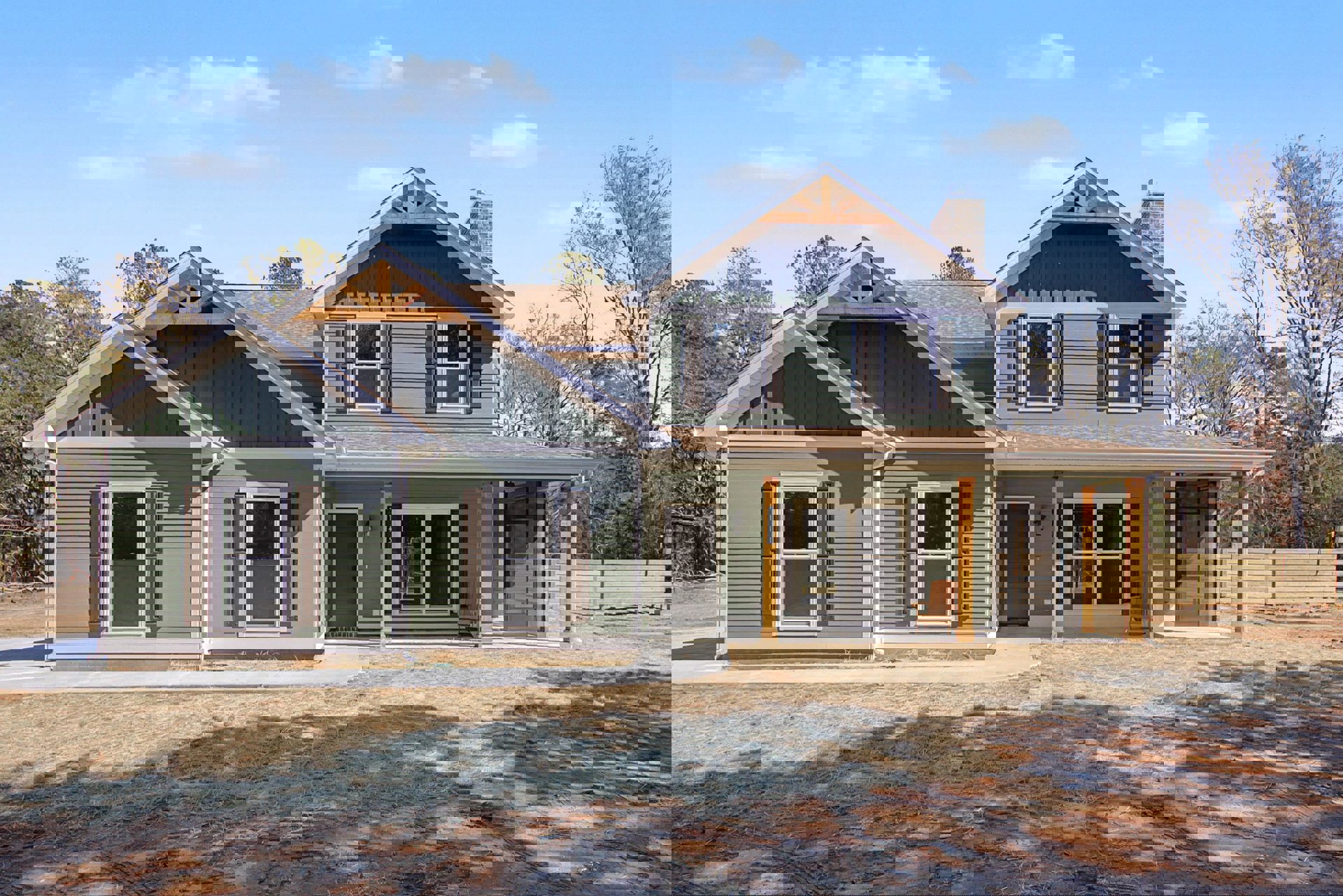 Two-story home with gray siding, gabled roof, paved driveway, covered front porch with wooden chair, manicured yard, mature trees, and large windows