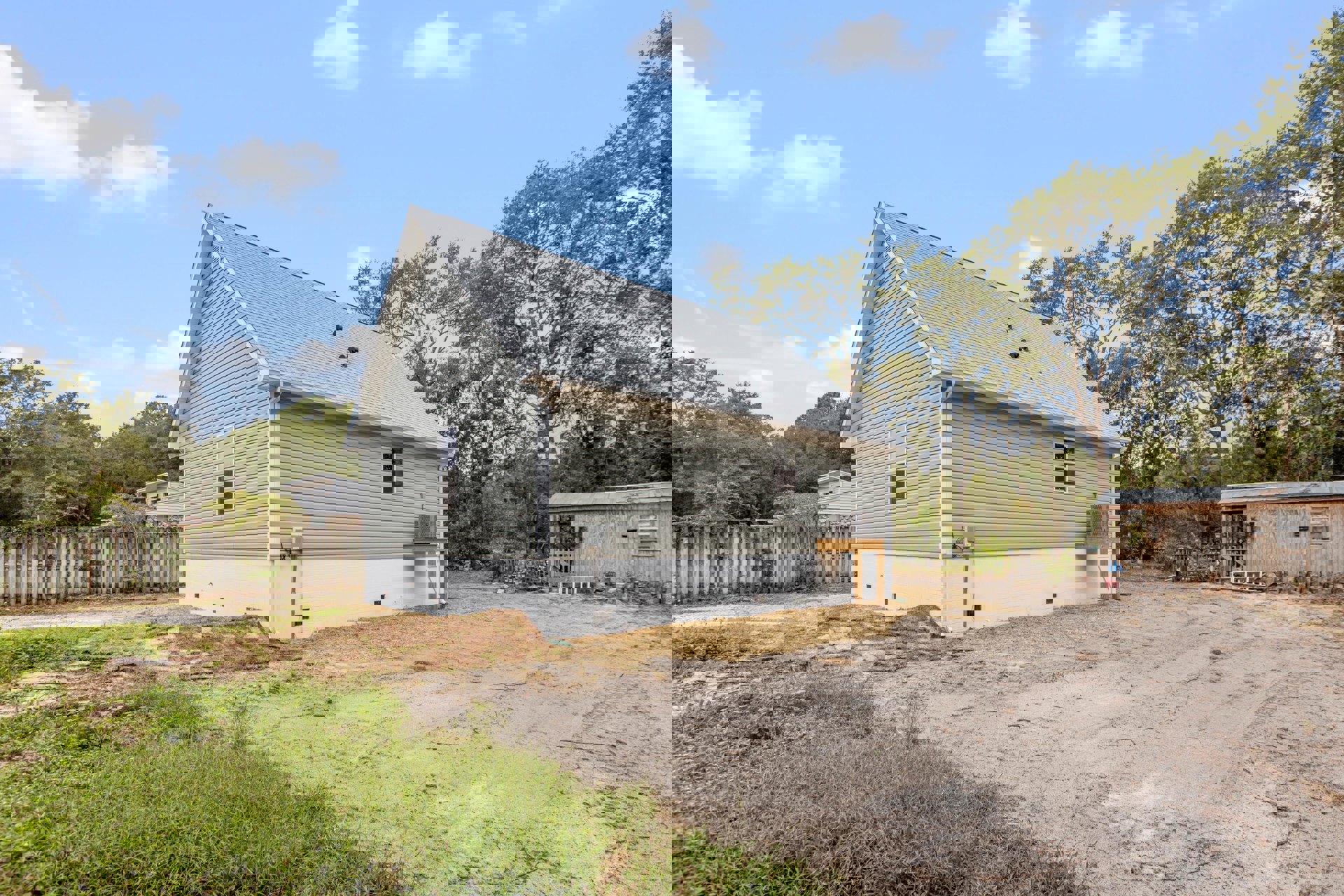 White cottage with gabled roof, wooden fence lined with climbing plants, dirt driveway bordered by grass, small shed and ladder visible in yard