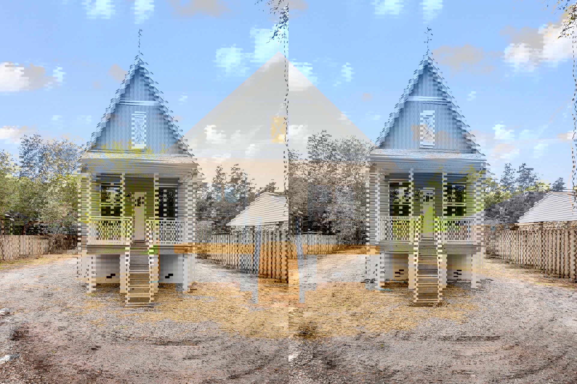 Two-story house with white siding, covered porch, wooden stairs, glass-paneled front door, wooden fence, and grassy yard with trees under a cloudy sky