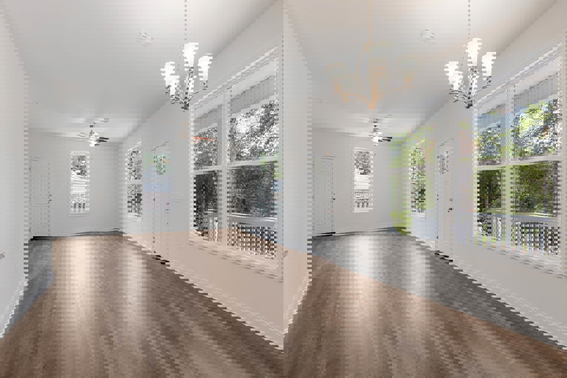 Wood flooring in a spacious room with a modern chandelier, white door with silver handle, large window framing chimney and trees outside, neutral walls and ceiling.