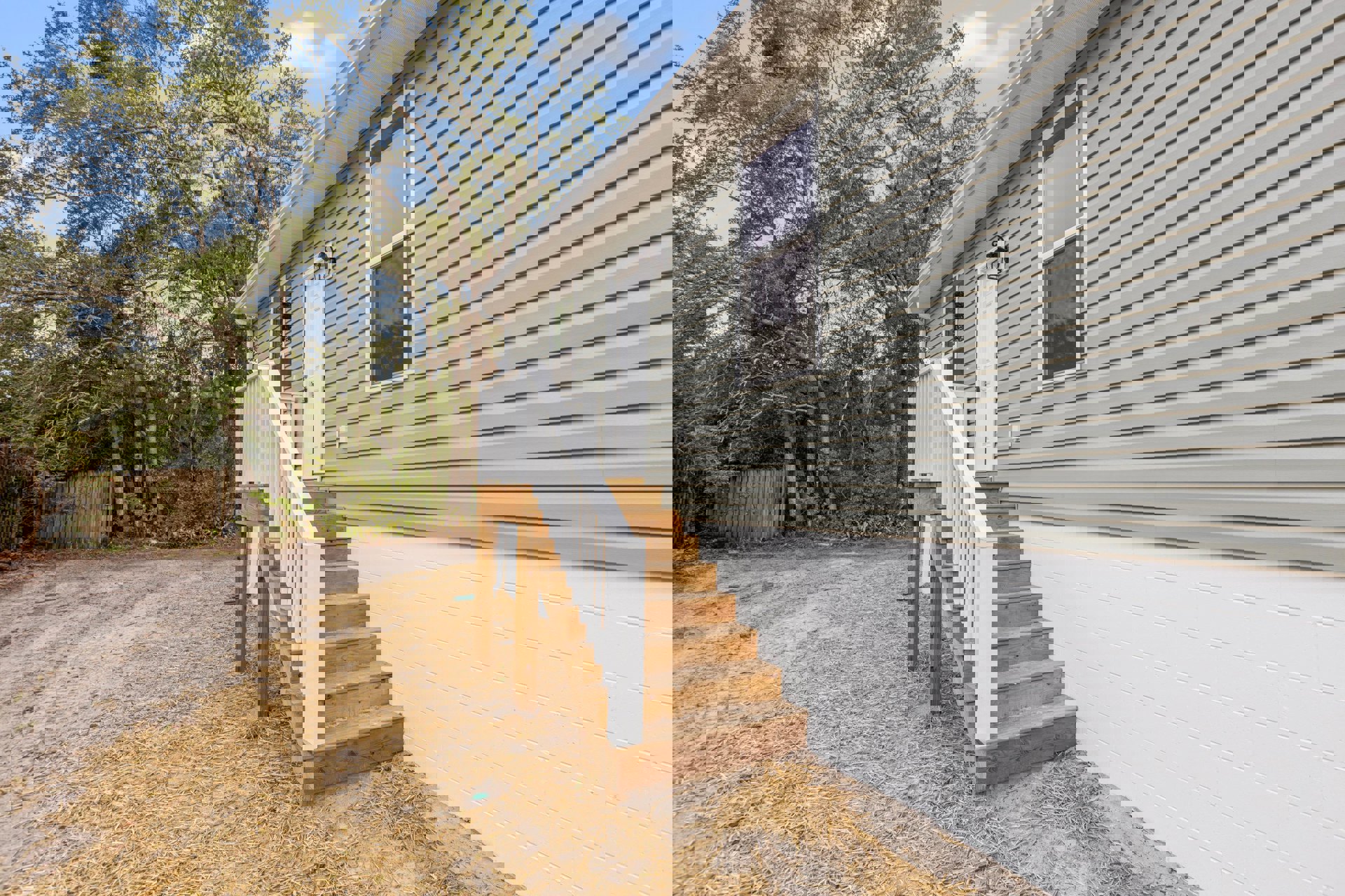 Two-story home with light siding, outdoor wooden staircase with white railing, white front door, screened window, and mature trees surrounding porch area