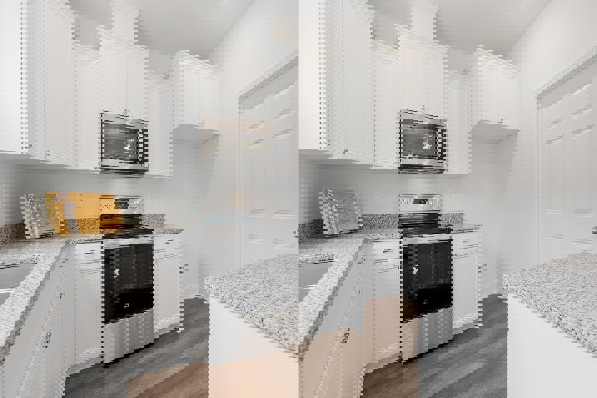 White shaker cabinets, black and silver oven, built-in microwave with illuminated interior, white countertop with black specks, wooden cutting board with ribbon bow.