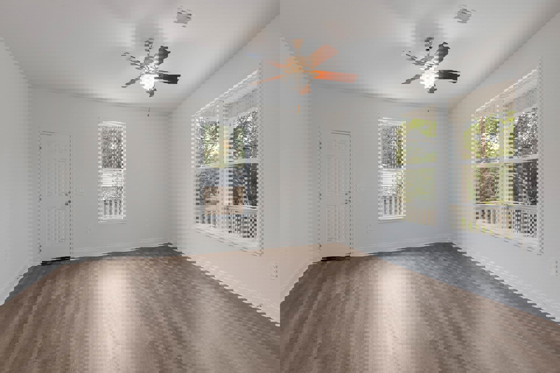 Living room with wood laminate flooring, white walls, ceiling fan with light fixture, large windows showing trees and a distant tower, white door with silver knob.