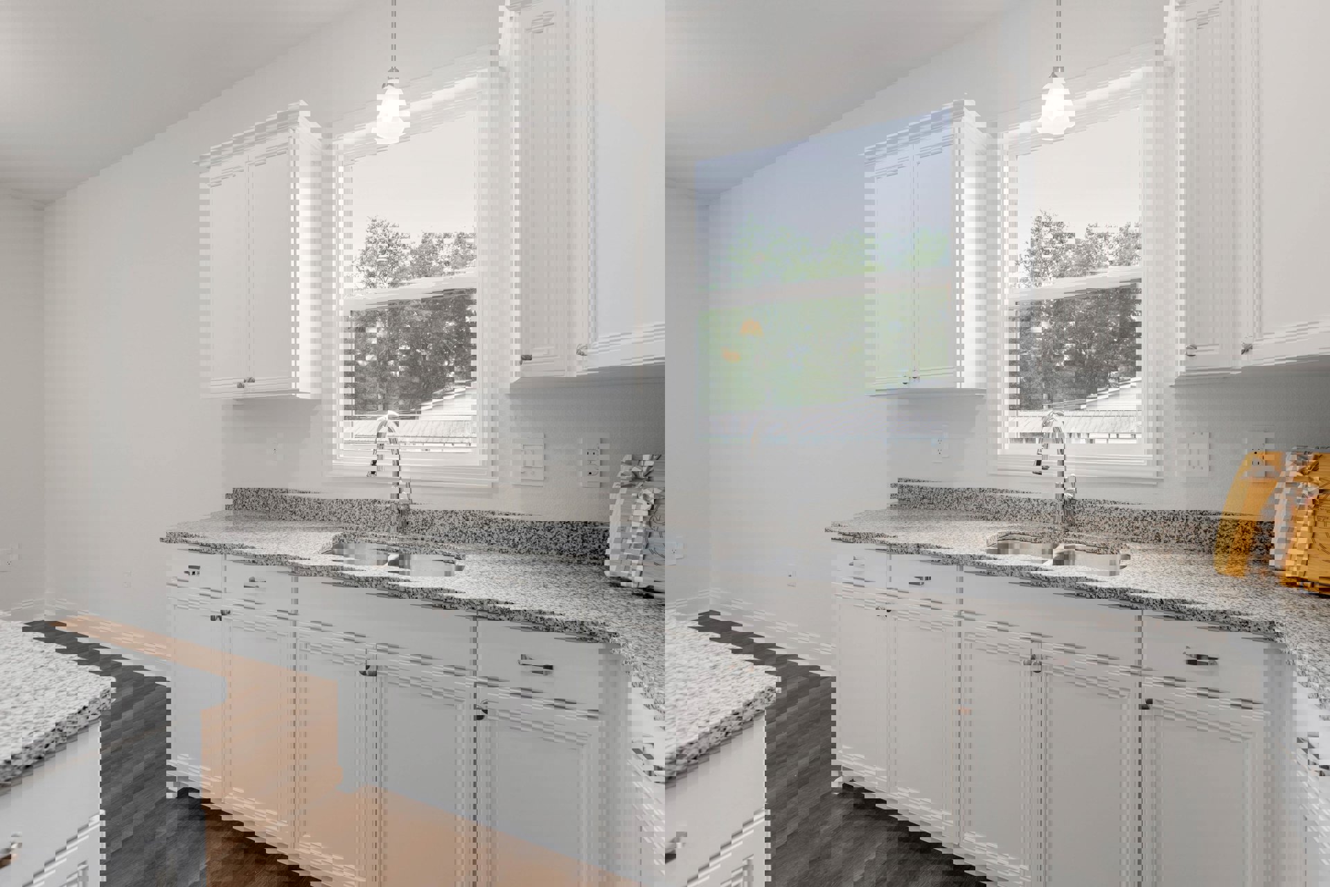 White kitchen cabinets and granite countertops, stainless steel sink beneath a window with tree views, wooden cutting board with ribbon on counter, white door and matching