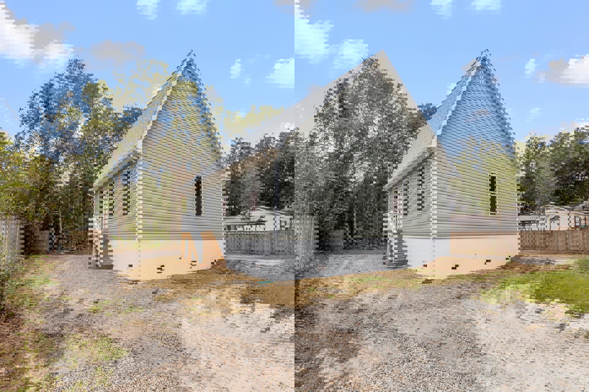 White house with grey roof, white brick walls, wooden exterior stairs, dirt path leading to entrance, tree branches overhead, blue sky with clouds, wooden fence along property.