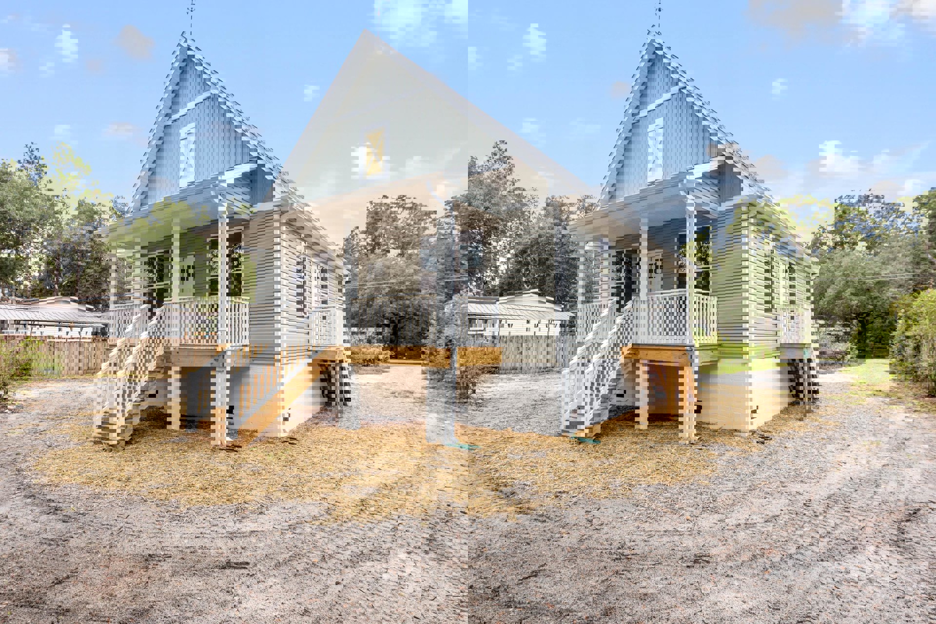 White brick house with wooden porch, white X-panel door, white stairs, straw mulch near entry, gabled roof, surrounded by trees and cloudy sky.