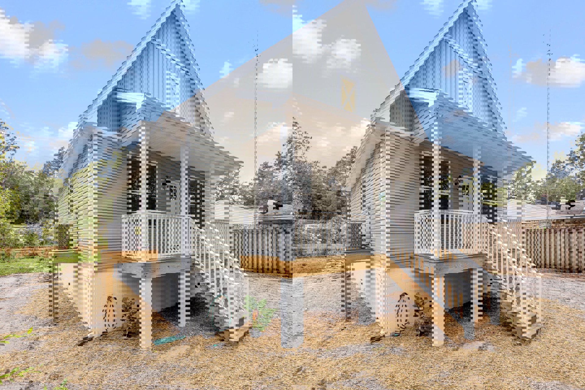 Two-story home with light siding, wooden porch and stairs, white front door, potted plant, garden hose mounted on wall, wooden fence and stone retaining wall, windows and trees