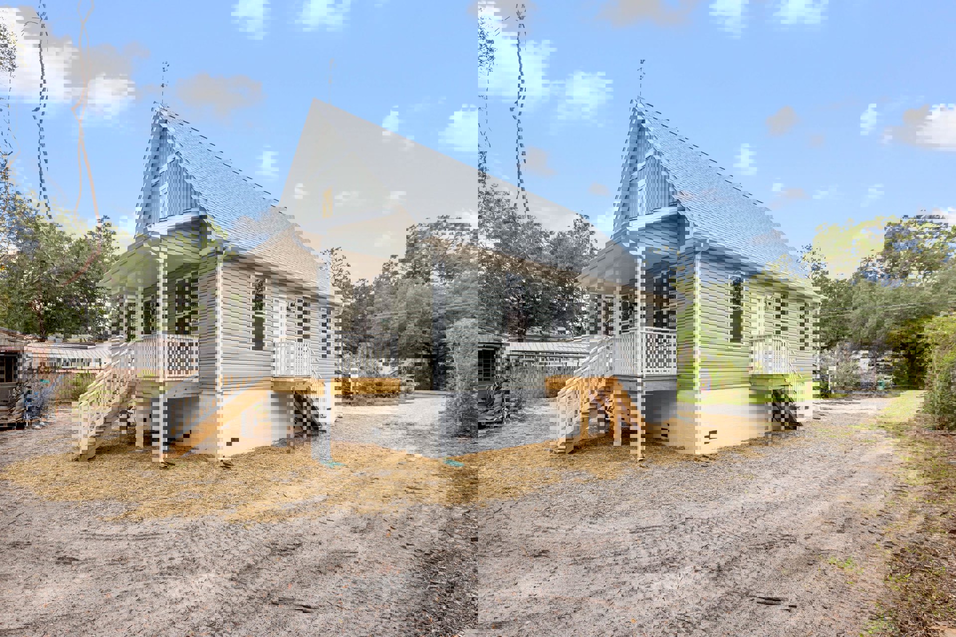 White two-story house with covered porch, white railing and staircase, wooden steps, dirt yard with tire tracks, scattered tools on porch, cloudy sky overhead.