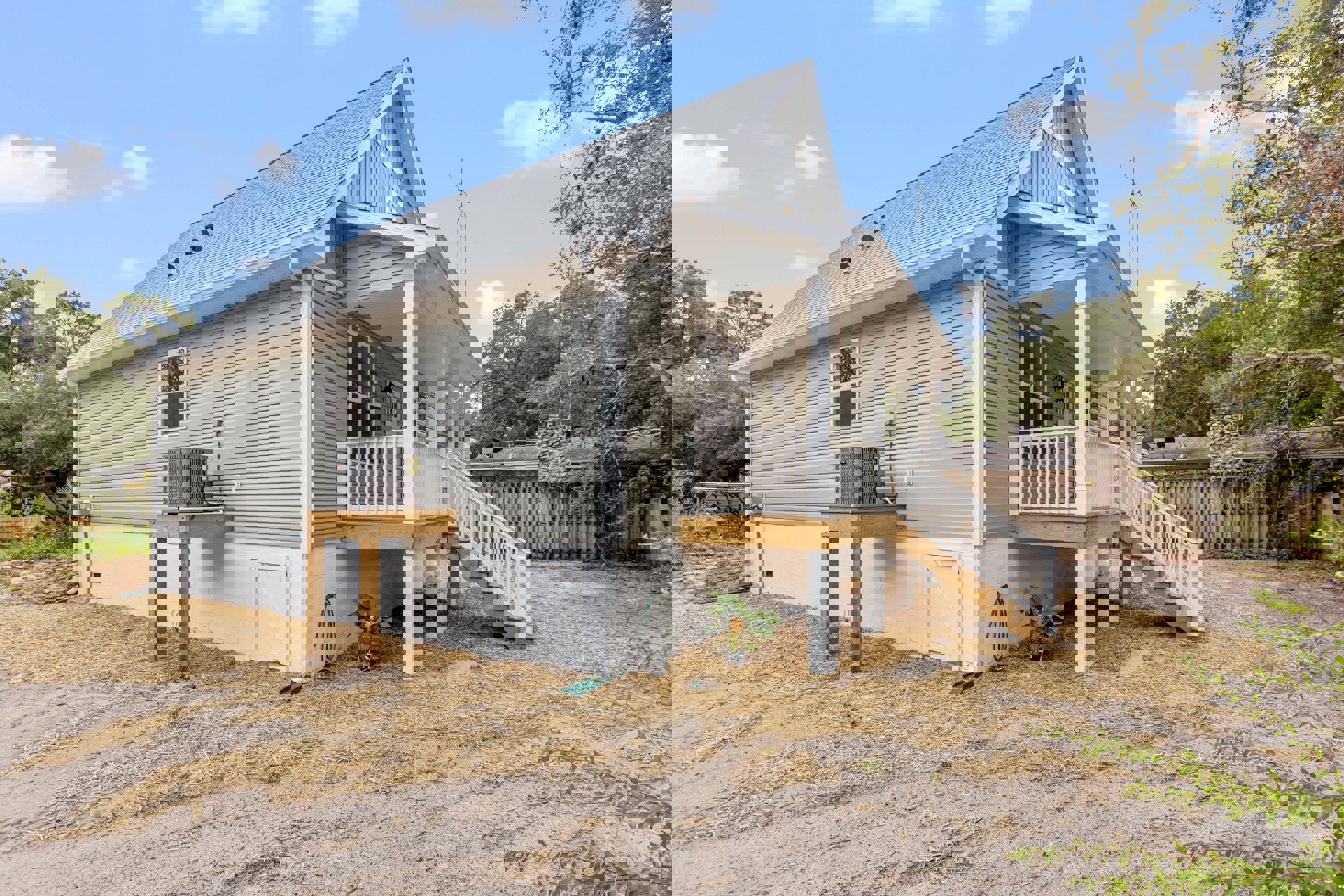 White cottage-style home with covered front porch, wooden railing, and picket fence; potted plant on porch, manicured lawn, and leafy trees in background under partly cloudy sky