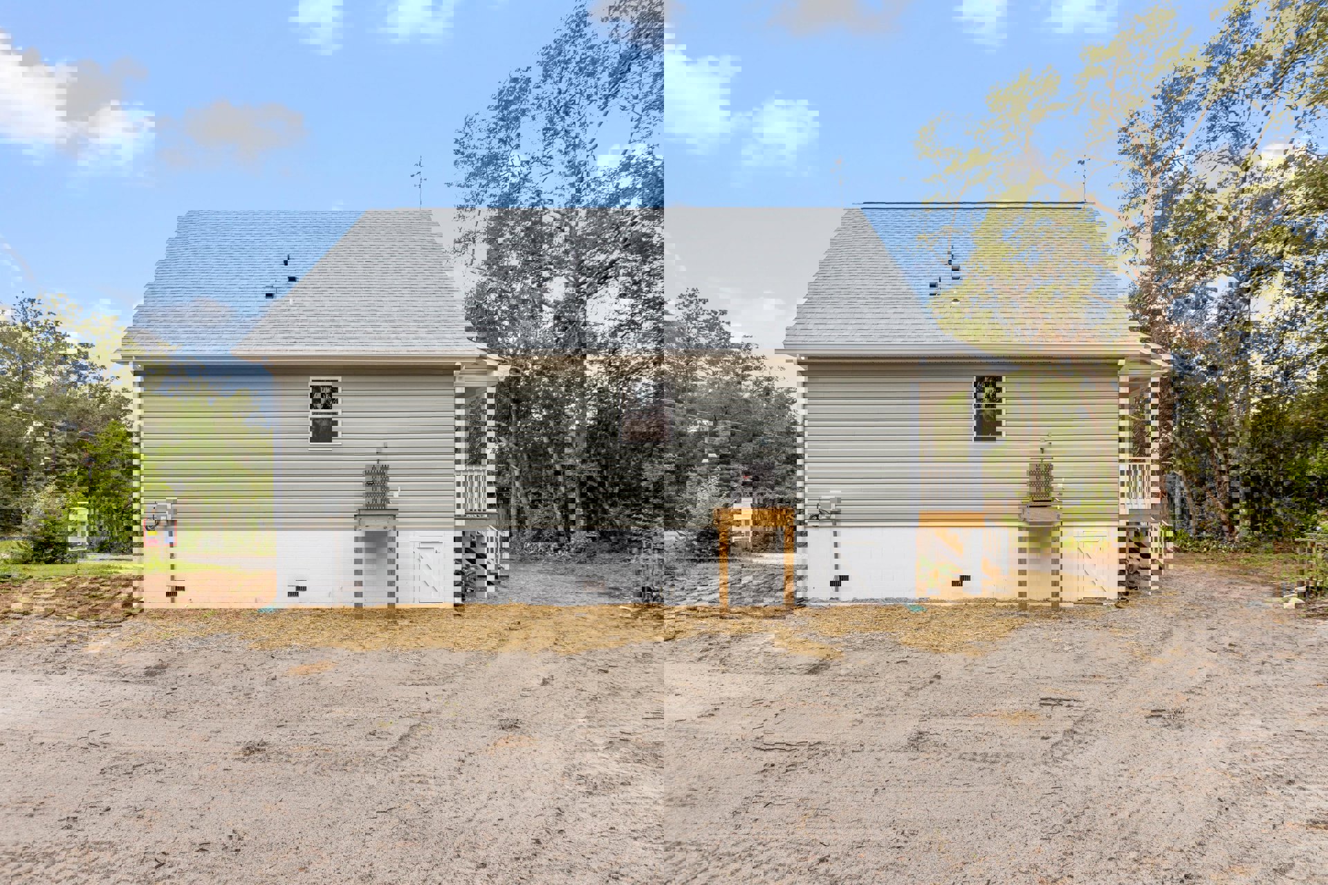Grey house with white-framed windows, wooden porch, and white picket fence; landscaped yard with trees and shrubs under a cloudy sky.
