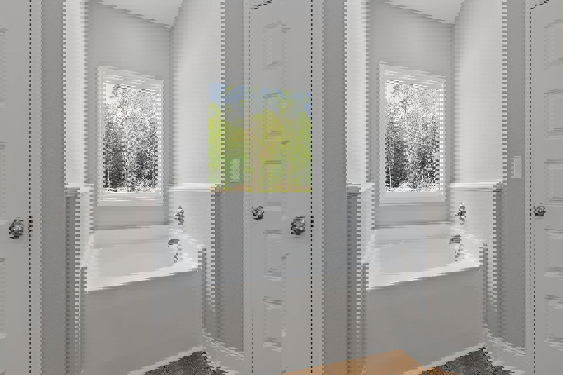 Freestanding white bathtub with silver faucets beneath a large window overlooking trees, light switch on adjacent wall, close-up of metal door knob.