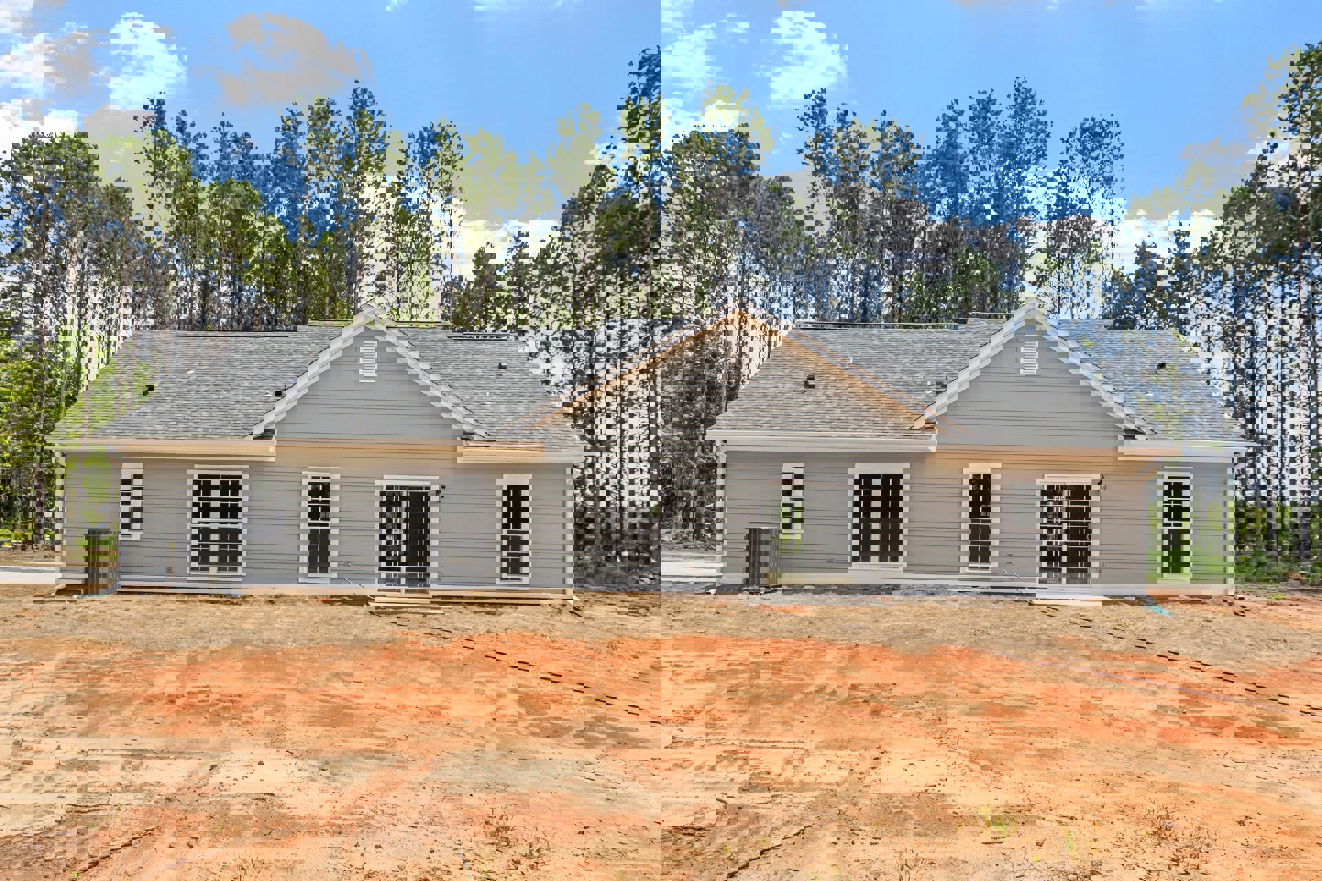 Partially built home with exposed framing, white-framed windows, sliding glass door, and garden hose attached to exterior wall; mature trees and small white cottage visible in