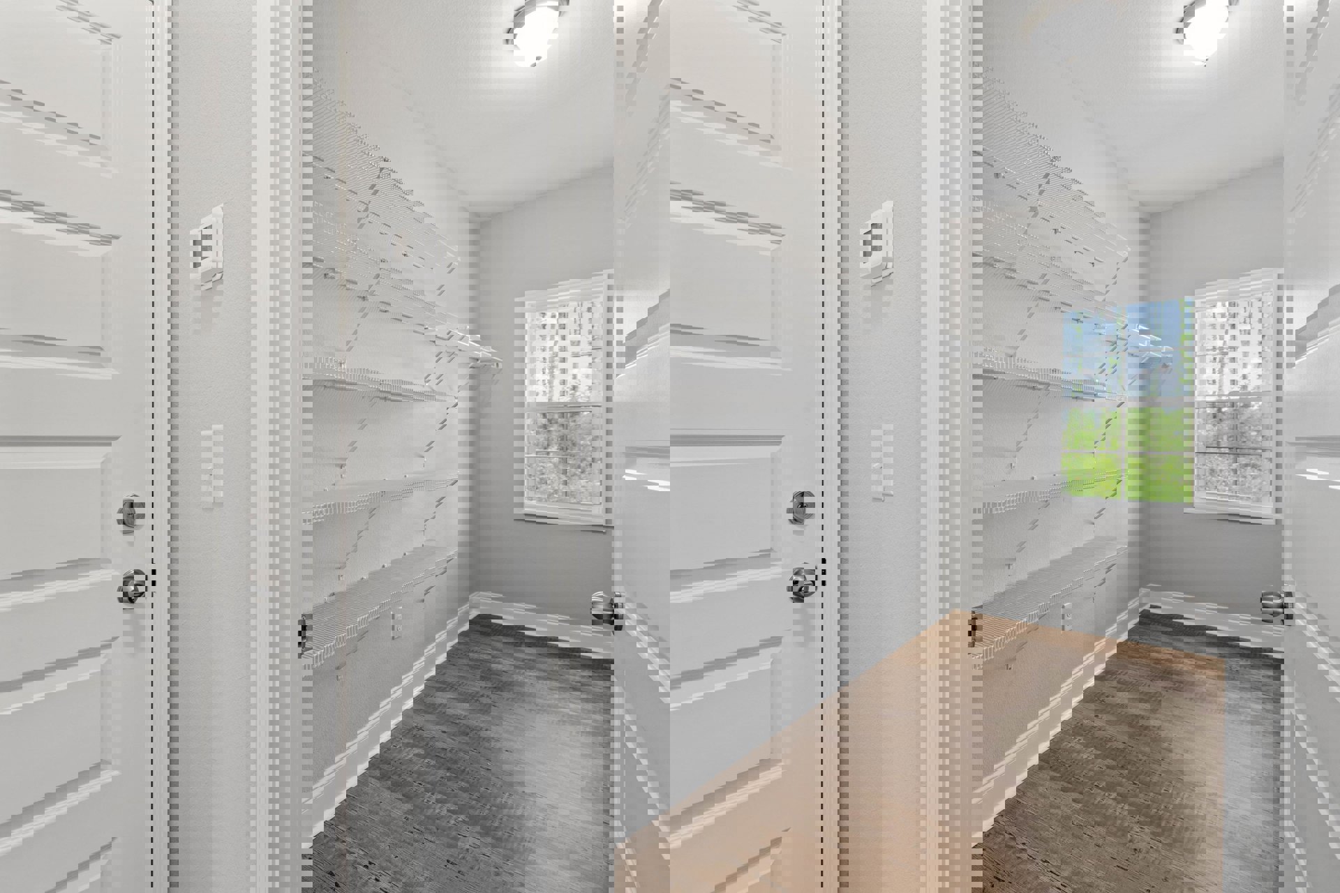 White built-in shelves against a plaster wall, wood floor with white baseboard trim, white door with silver knobs, window overlooking green trees, silver decorative object on shelf