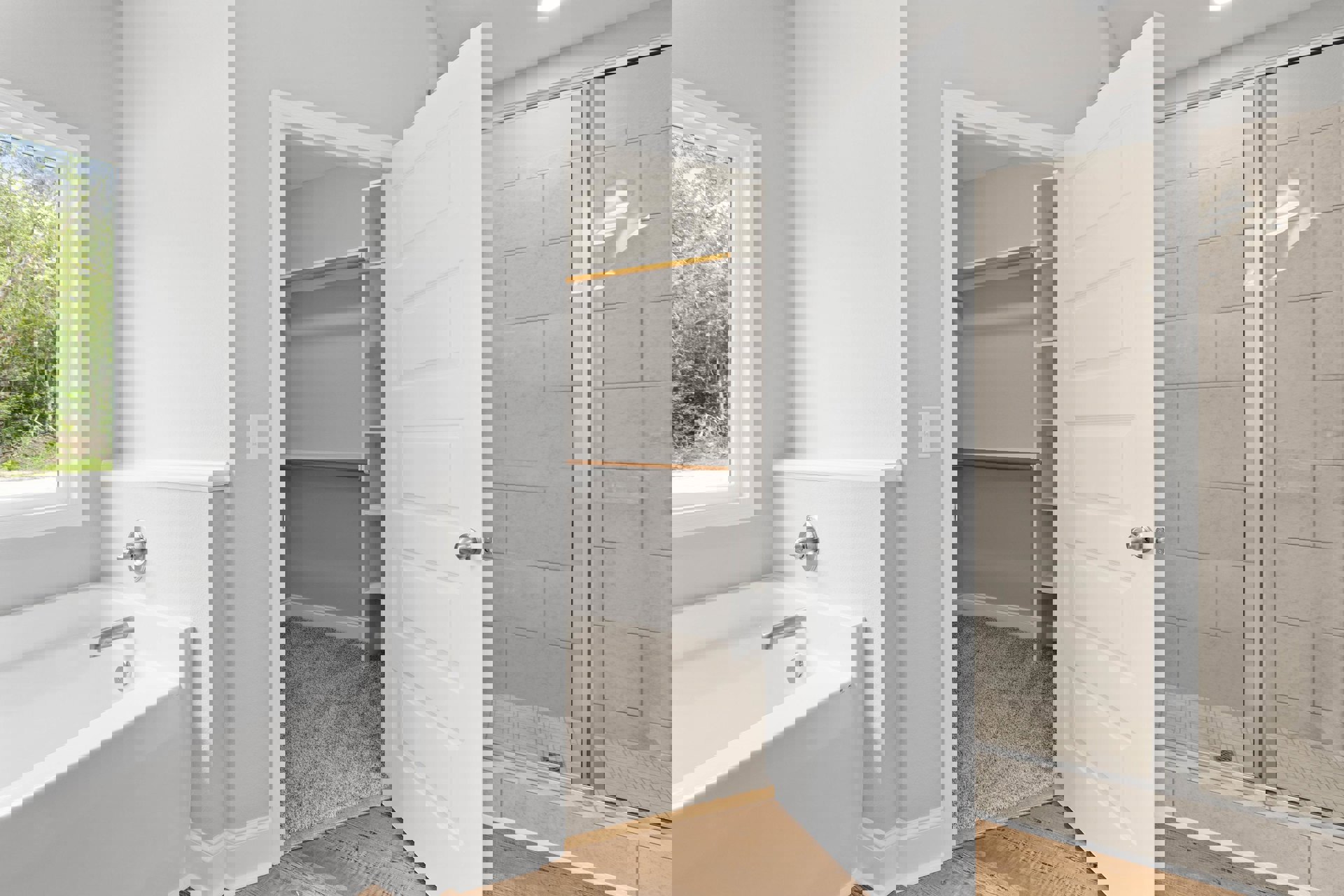 Walk-in closet adjacent to bathroom with white bathtub and silver faucet, glass shower door on tiled wall, window overlooking trees, white door with silver knob