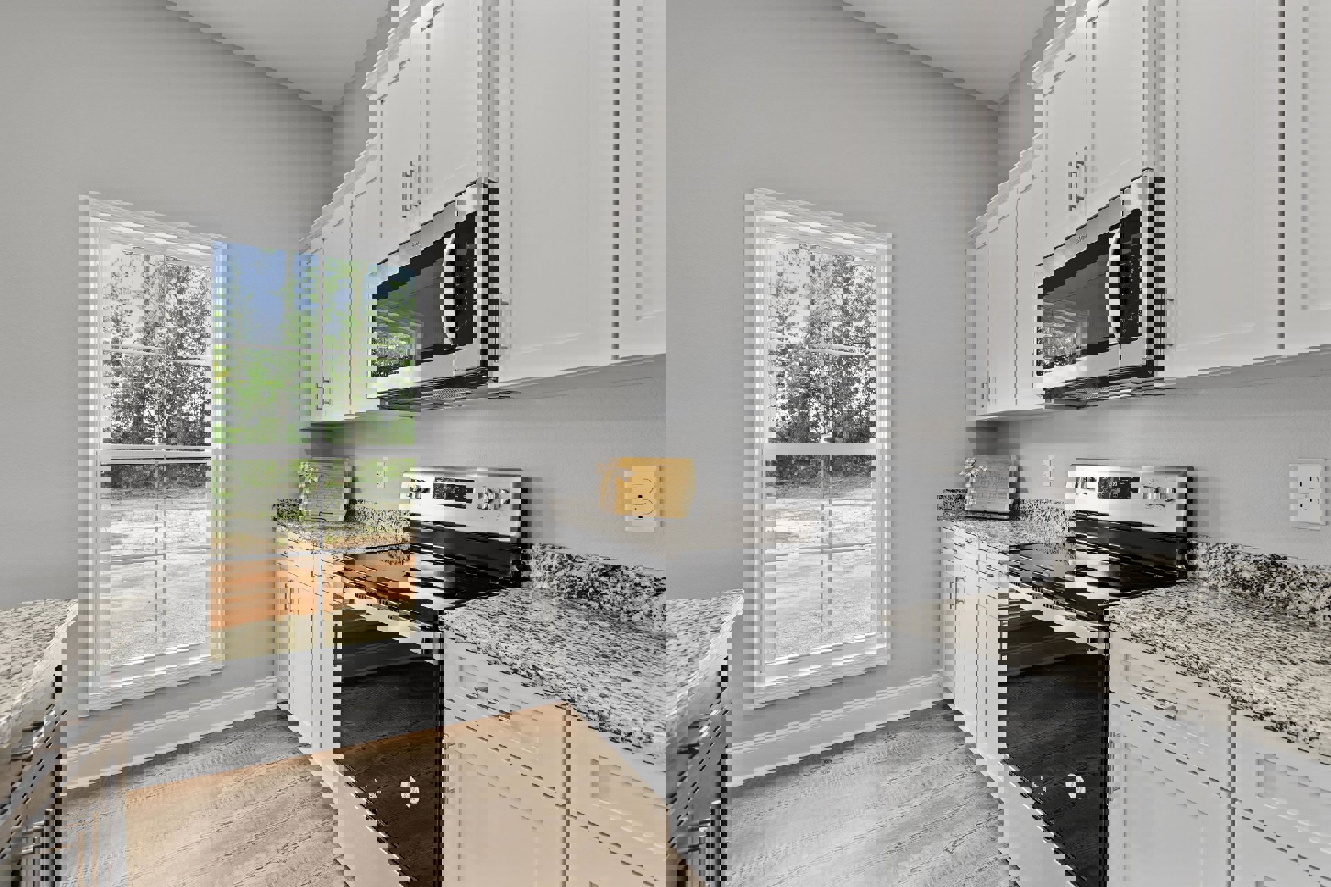 White shaker cabinets, black freestanding stove, built-in microwave, quartz countertops, window overlooking trees, electrical outlet on backsplash, wooden gift box with bow on