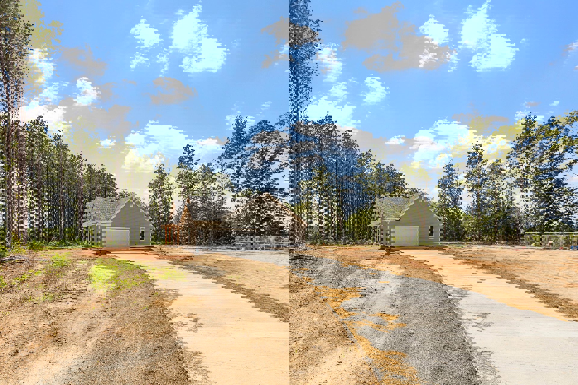 Two-story custom home with light siding, prominent garage door, concrete driveway, manicured lawn, and mature trees under a partly cloudy blue sky