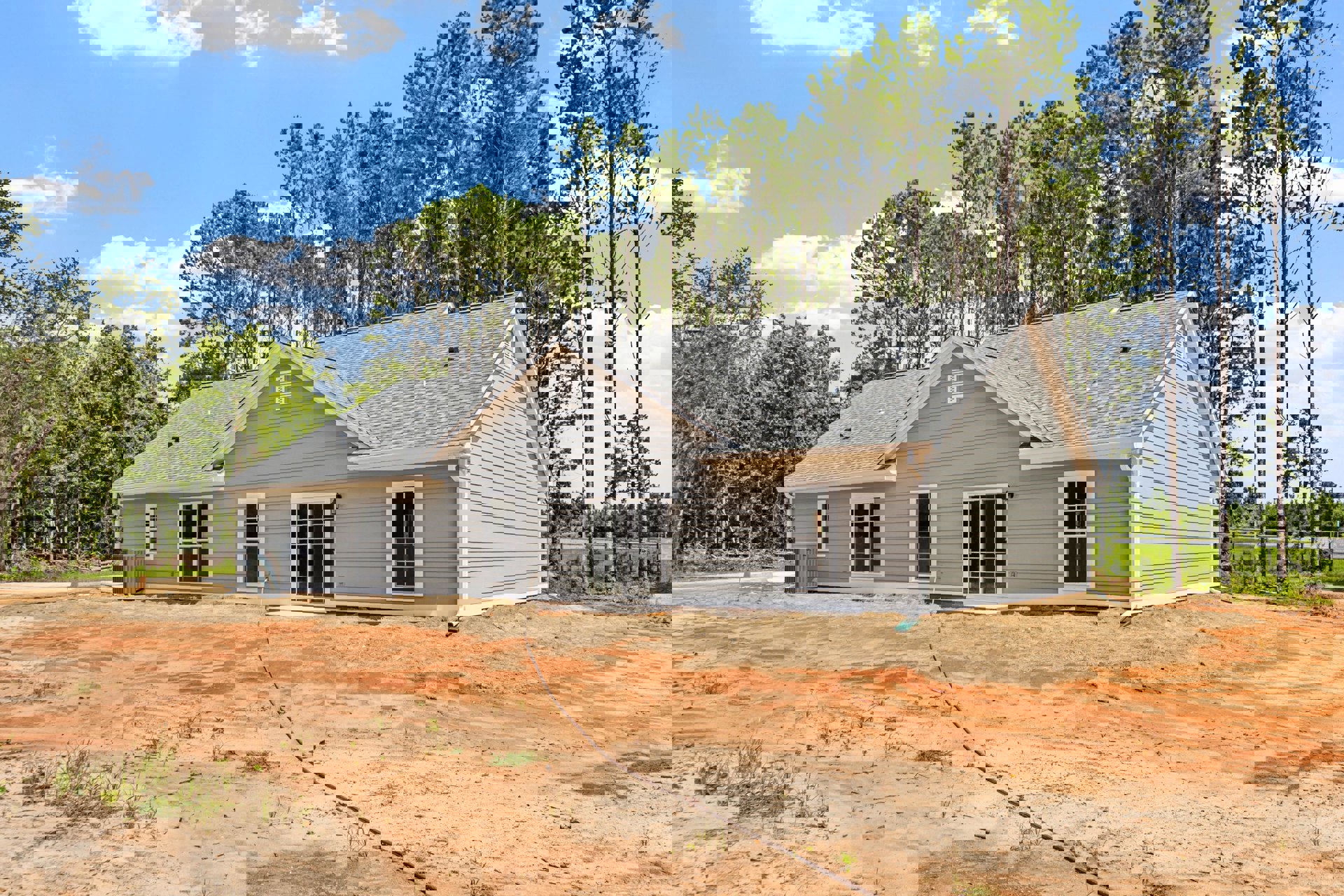 Partially built house with white-framed window, shingled roof, exposed dirt lot, long black hose, and trees in the background