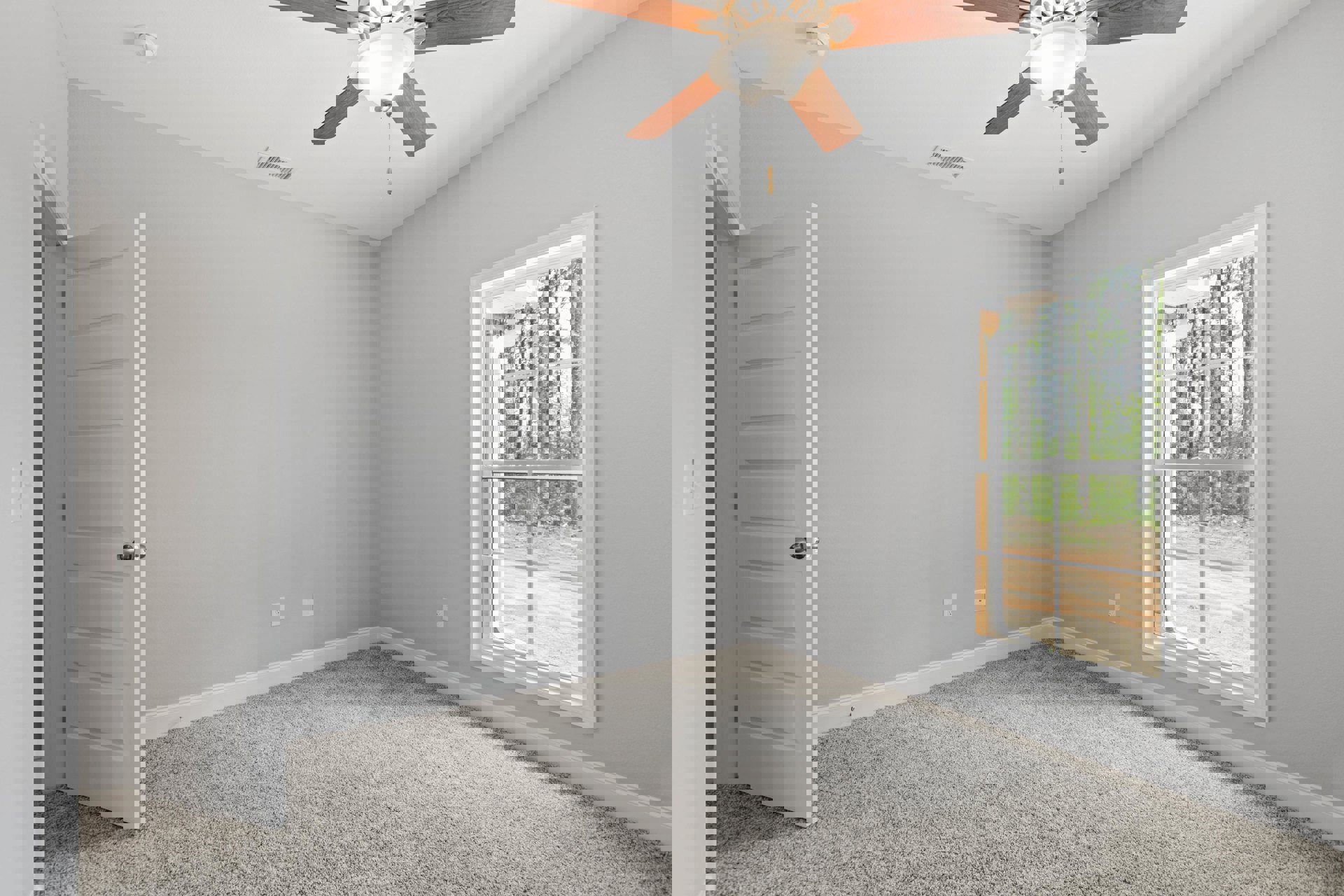 Carpeted room with white walls, ceiling fan with light fixture, large window showing trees and a dirt road, white door with silver knob, simple baseboard molding