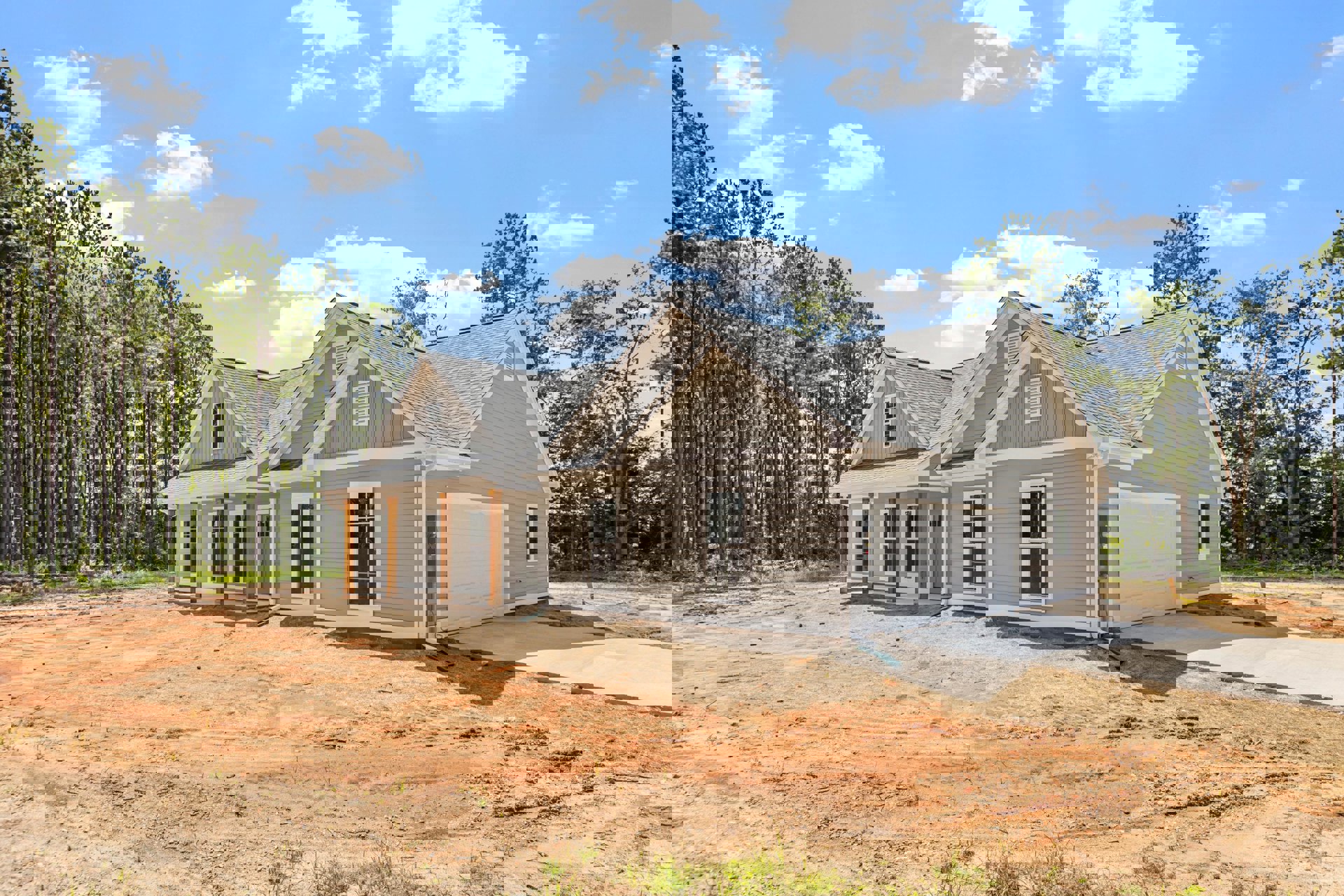 Partially built home with white framed window, exposed roof, dirt patch in front, garage on the side, surrounded by trees and blue sky with clouds; Little White House visible in