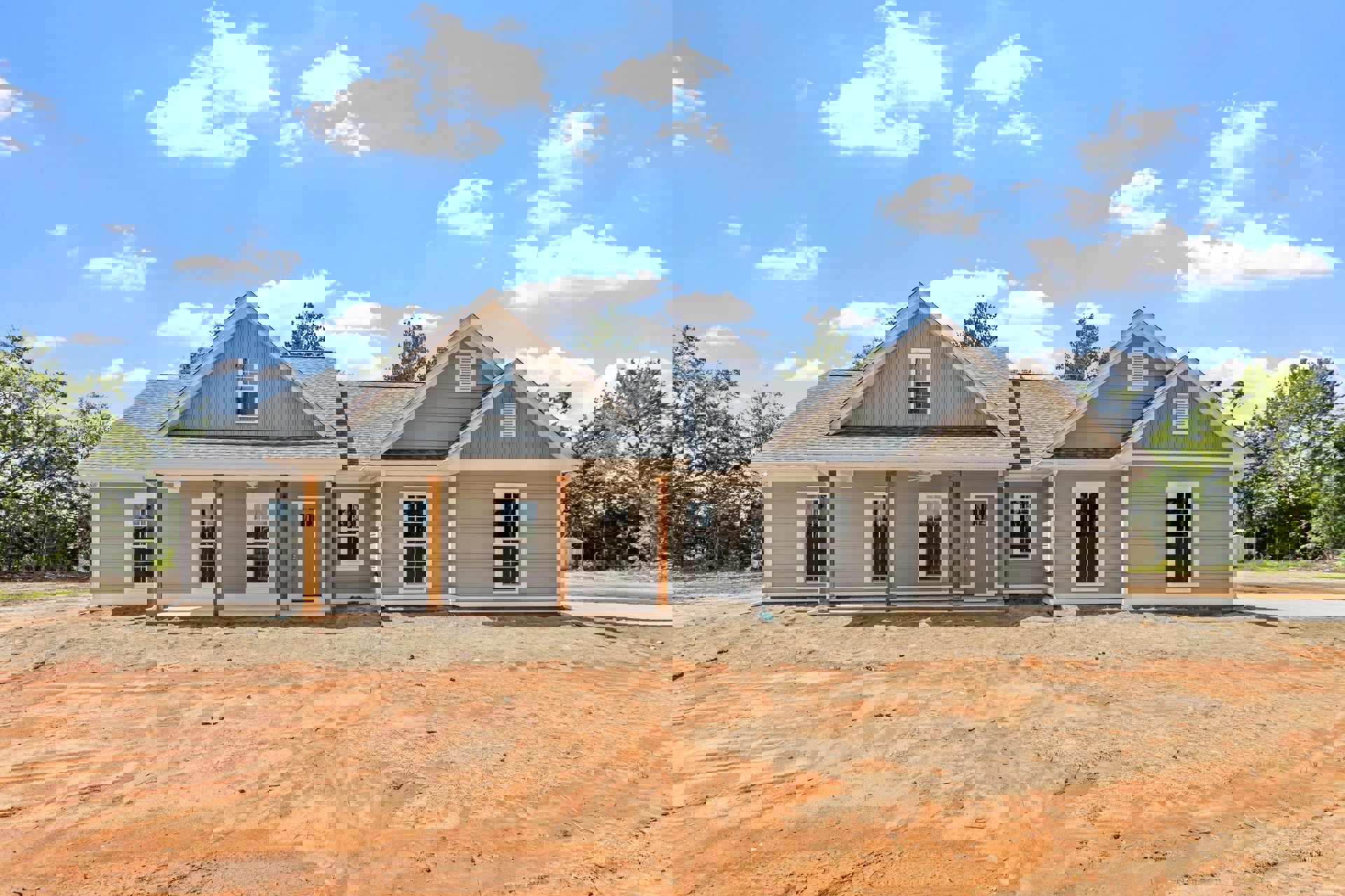 Two-story house under construction with exposed wood framing, white window frames, covered porch, dirt yard, and blue sky with scattered clouds