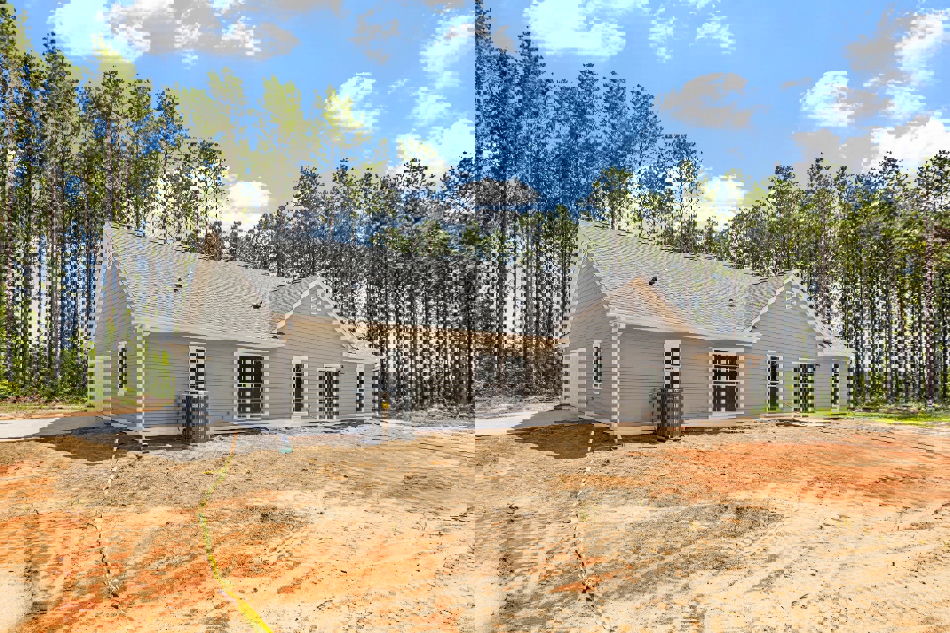 Partially built house with exposed framing, dirt yard, heat pump unit near exterior wall, large windows, and mature trees in background