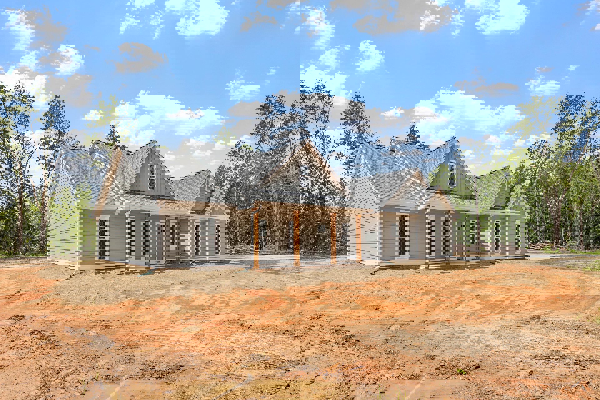 Partially built house with exposed framing, dirt yard, surrounding trees, and small white cottage visible in the background under a blue sky with scattered clouds