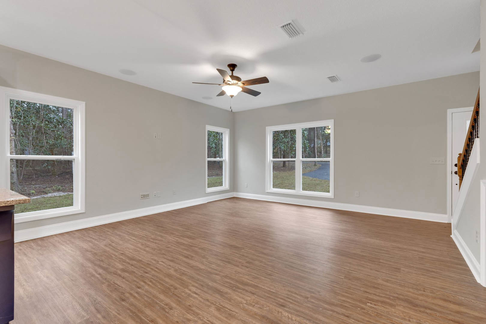 Room with hardwood flooring, ceiling fan with light fixture, large windows showing forest views, white walls, and a close-up of a wooden door