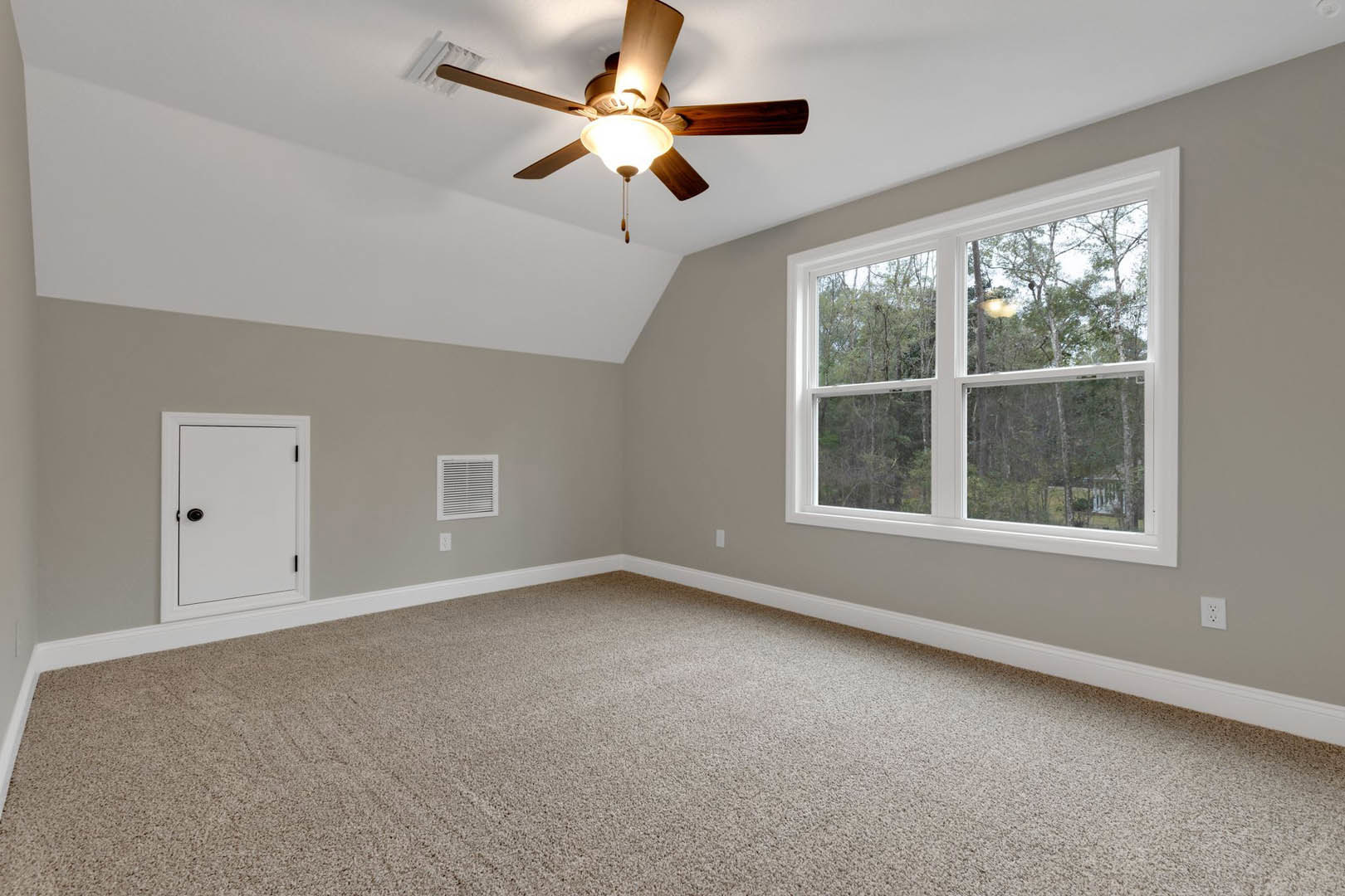 Carpeted room with white walls, ceiling fan with light, white door with black handle, window showing trees outside, wall vent, simple molding
