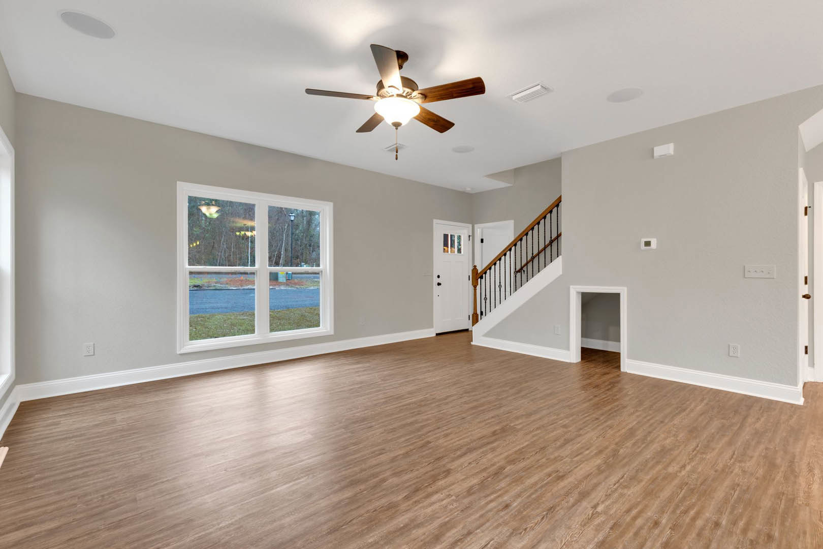 Wood flooring and white walls in a bright room with a ceiling fan and light fixture, window overlooking the street, staircase with wooden steps, and a white door with a sign.