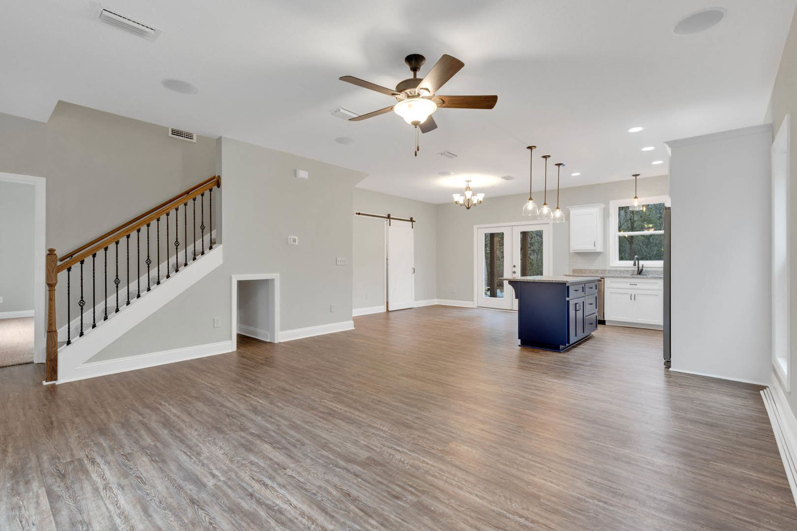 Spacious open-concept living area featuring wood flooring, blue kitchen island with marble countertop, modern ceiling fan with light, and a staircase with close-up detail; adjacent