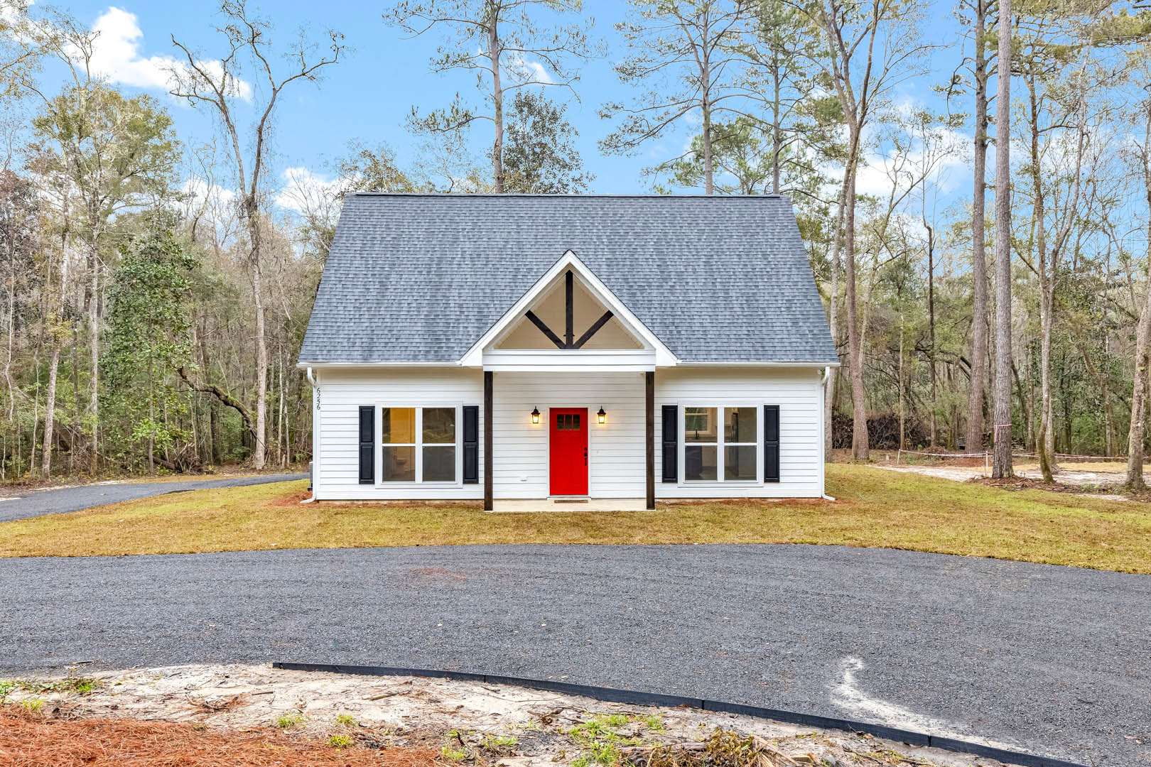 White siding house with red front door, paved driveway, green lawn, mature trees in background, large windows, partly cloudy sky