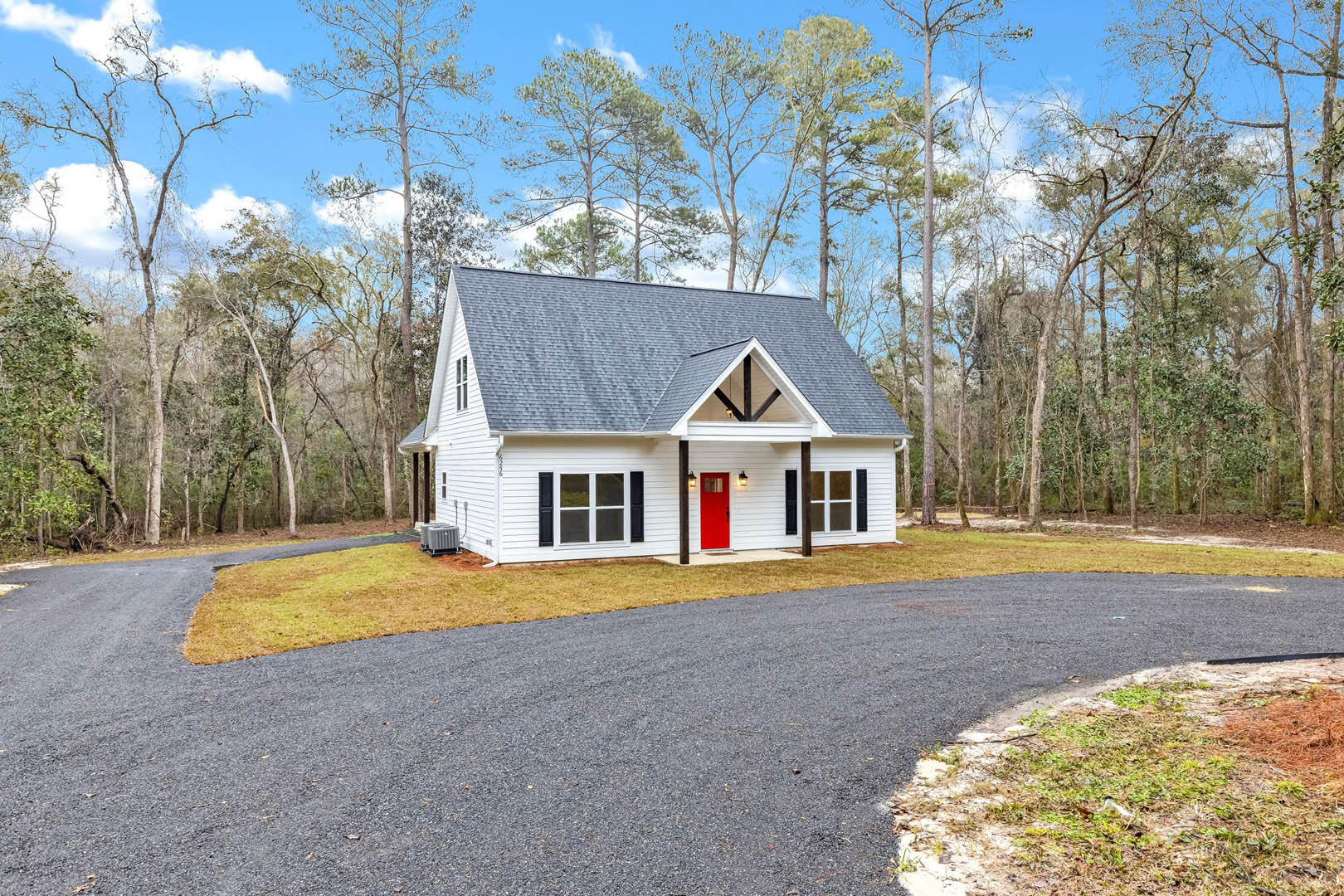 White house with red front door, paved driveway, green lawn, mature trees in background, cloudy sky overhead