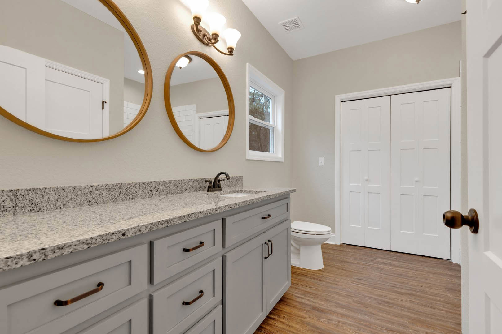Bathroom featuring marble countertop with undermount sink, white toilet, wood flooring, wall mirror, white closet doors, brown interior door, and window overlooking trees.