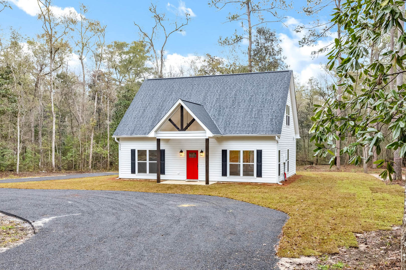 White siding house with a red front door, black framed windows, gabled roof, green leafy tree in front yard, paved road with yellow center line